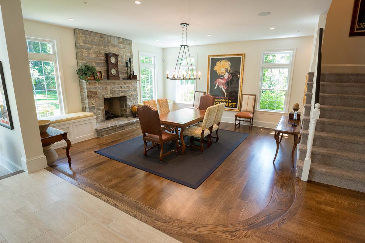 Warm dining room with wooden table, upholstered chairs, stone fireplace, vintage chandelier, and large windows near a staircase.