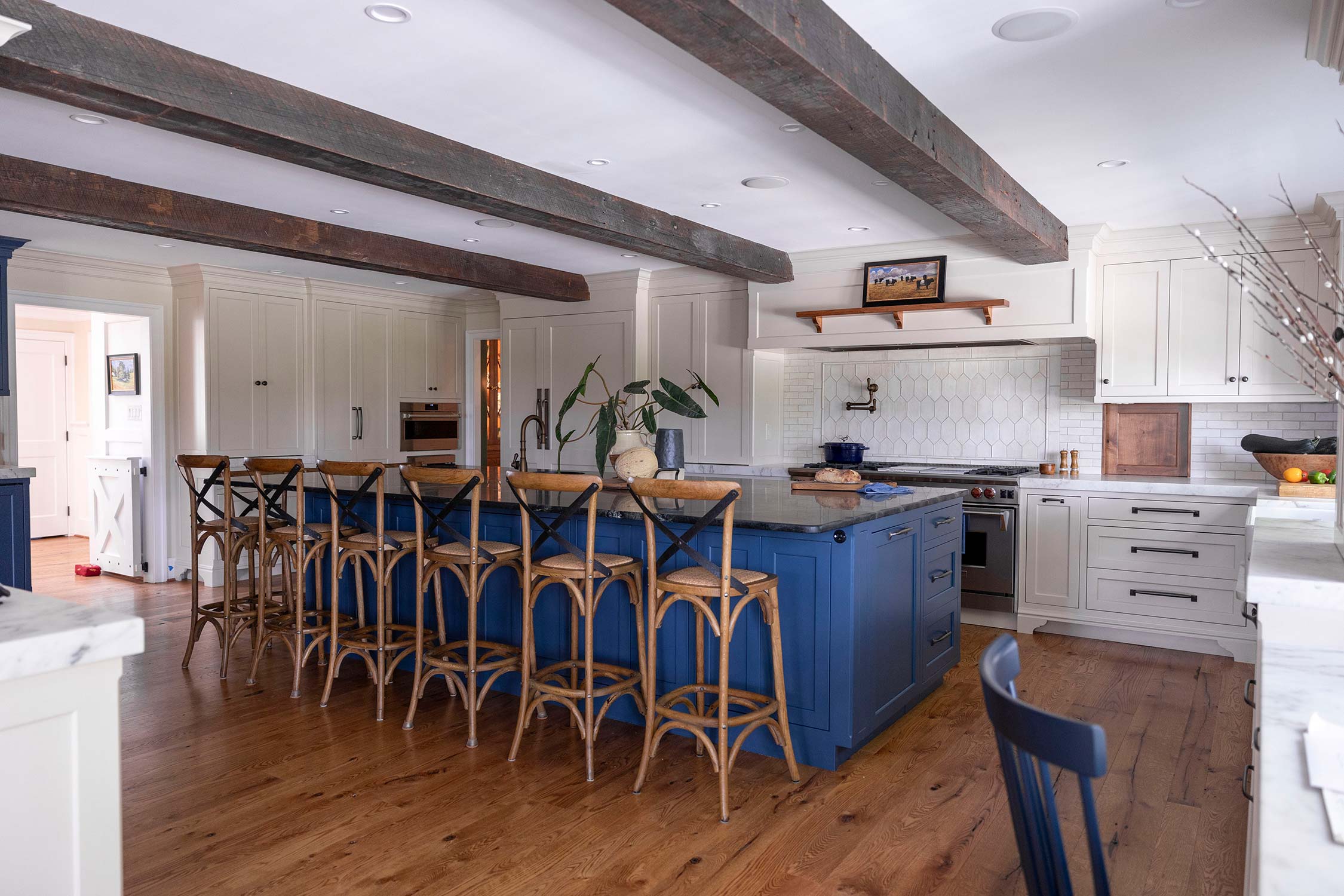 Spacious kitchen with blue island, wooden stools, white cabinets, marble countertops, and exposed wooden beams on the ceiling.