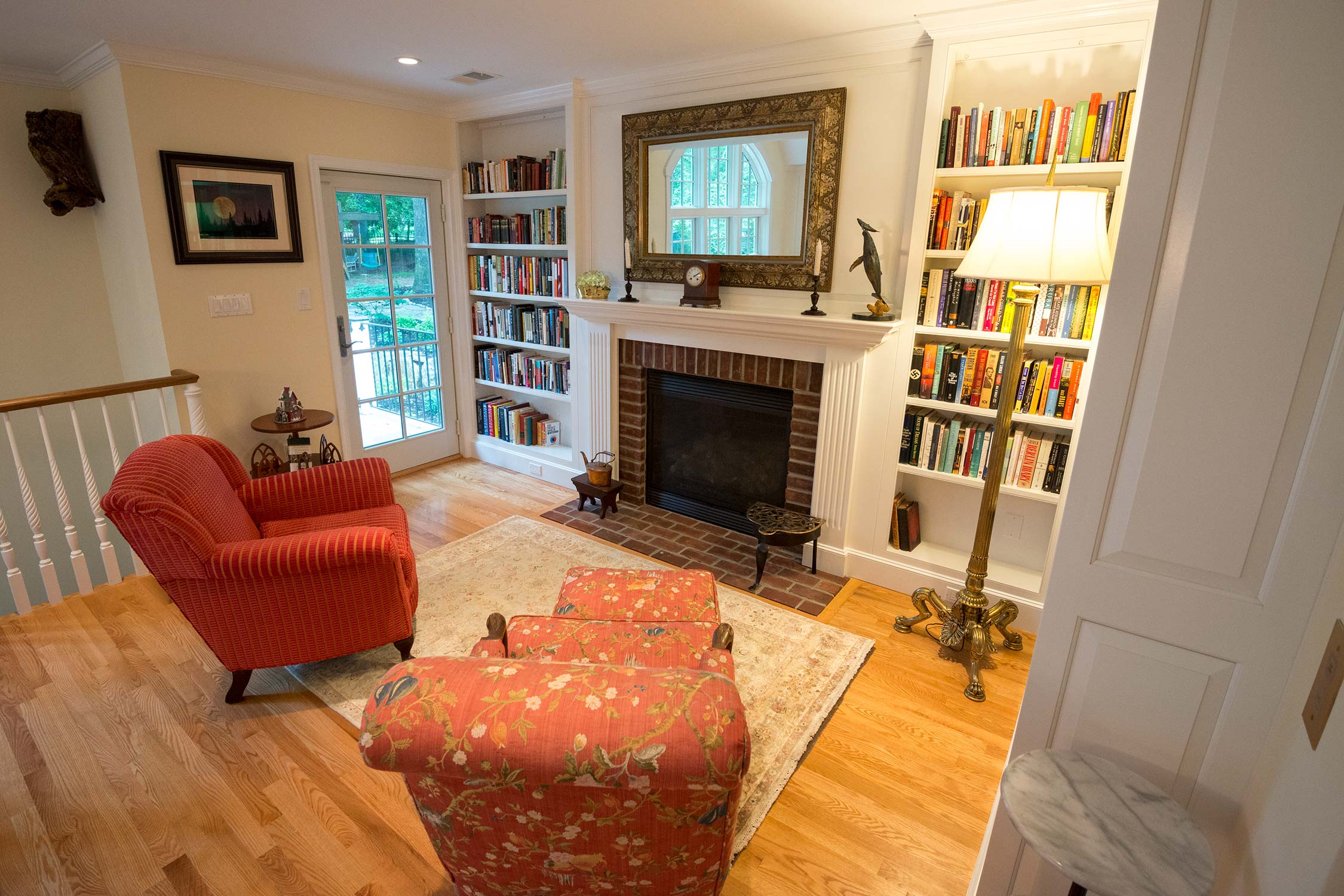 Cozy room with red floral armchairs, fireplace, and bookshelves. Features hardwood floors, a large mirror, and garden view through a glass door.