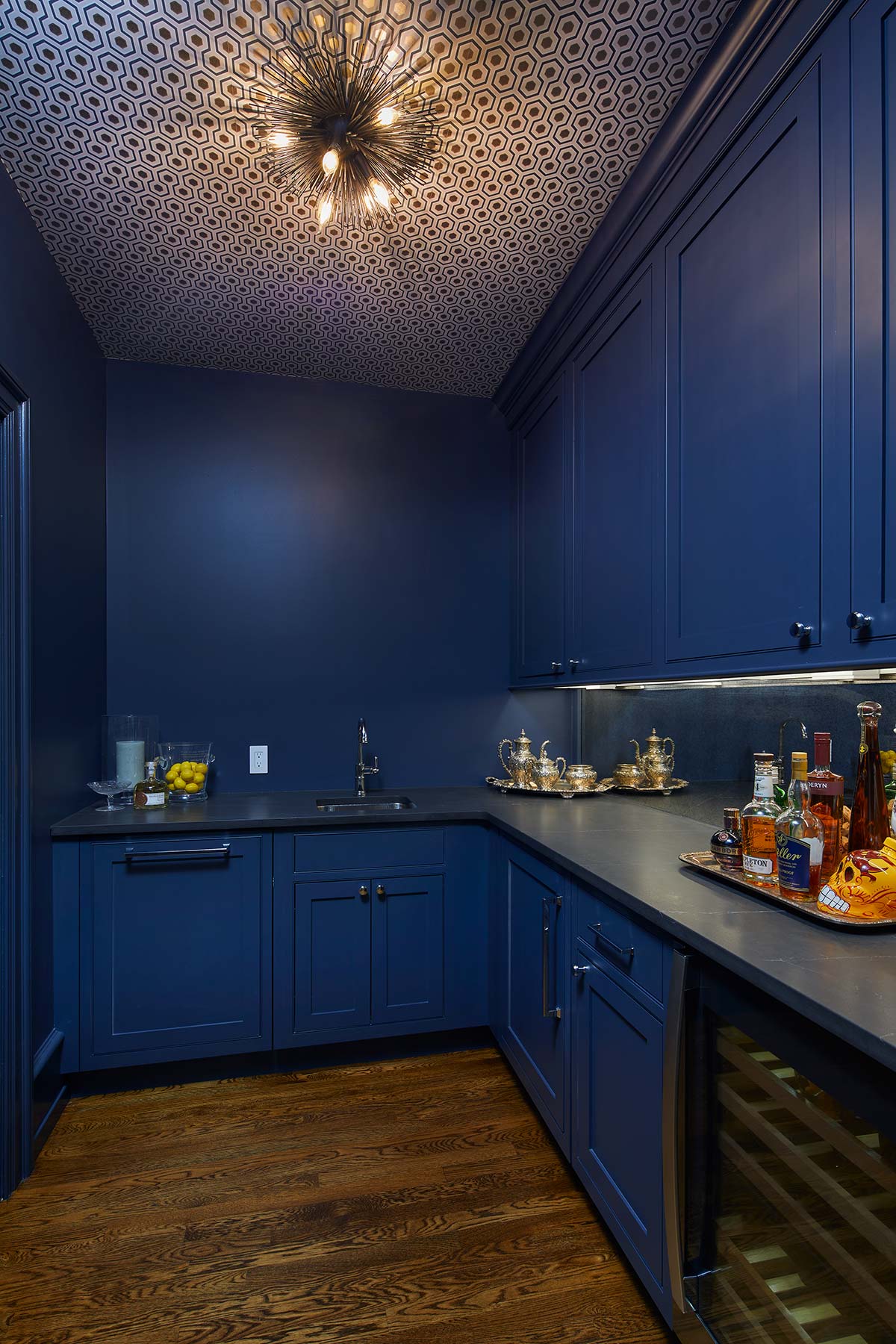 A stylish butler's pantry with navy cabinets, a patterned ceiling, modern chandelier, dark countertops, a sink, and a tray of liquor bottles and teapots.