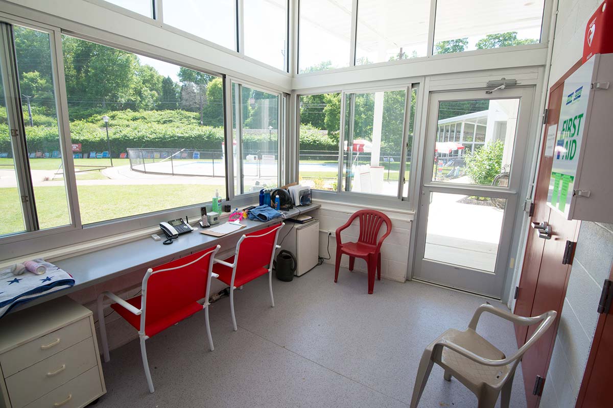 Lifeguard staff office with a desk, red chairs, phone, and supplies, surrounded by large windows. A first aid kit is on the wall, and a pool area is visible outside.