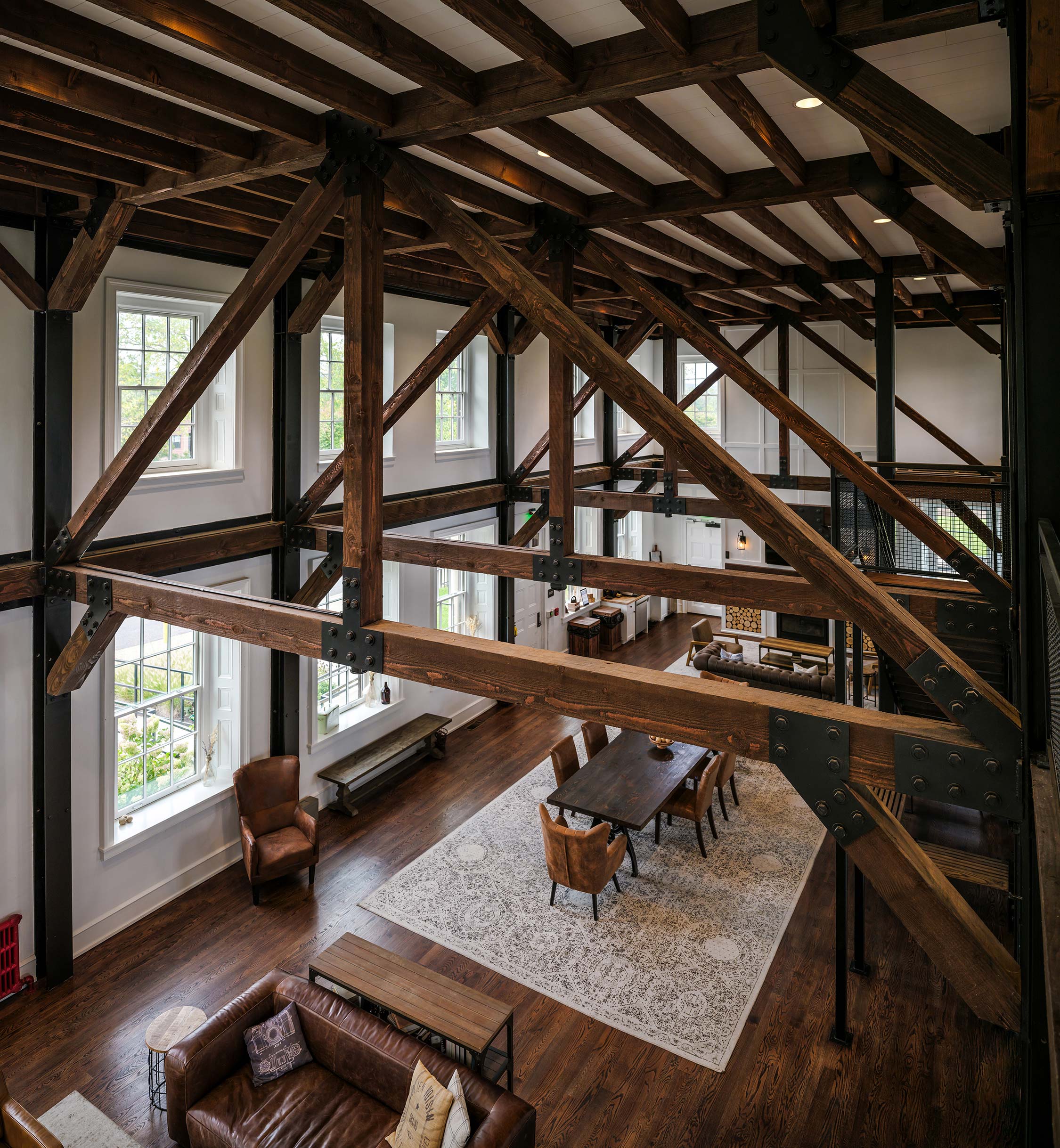 Open room with wooden beams, large windows, dining table, leather chairs, and sofa on a patterned rug.