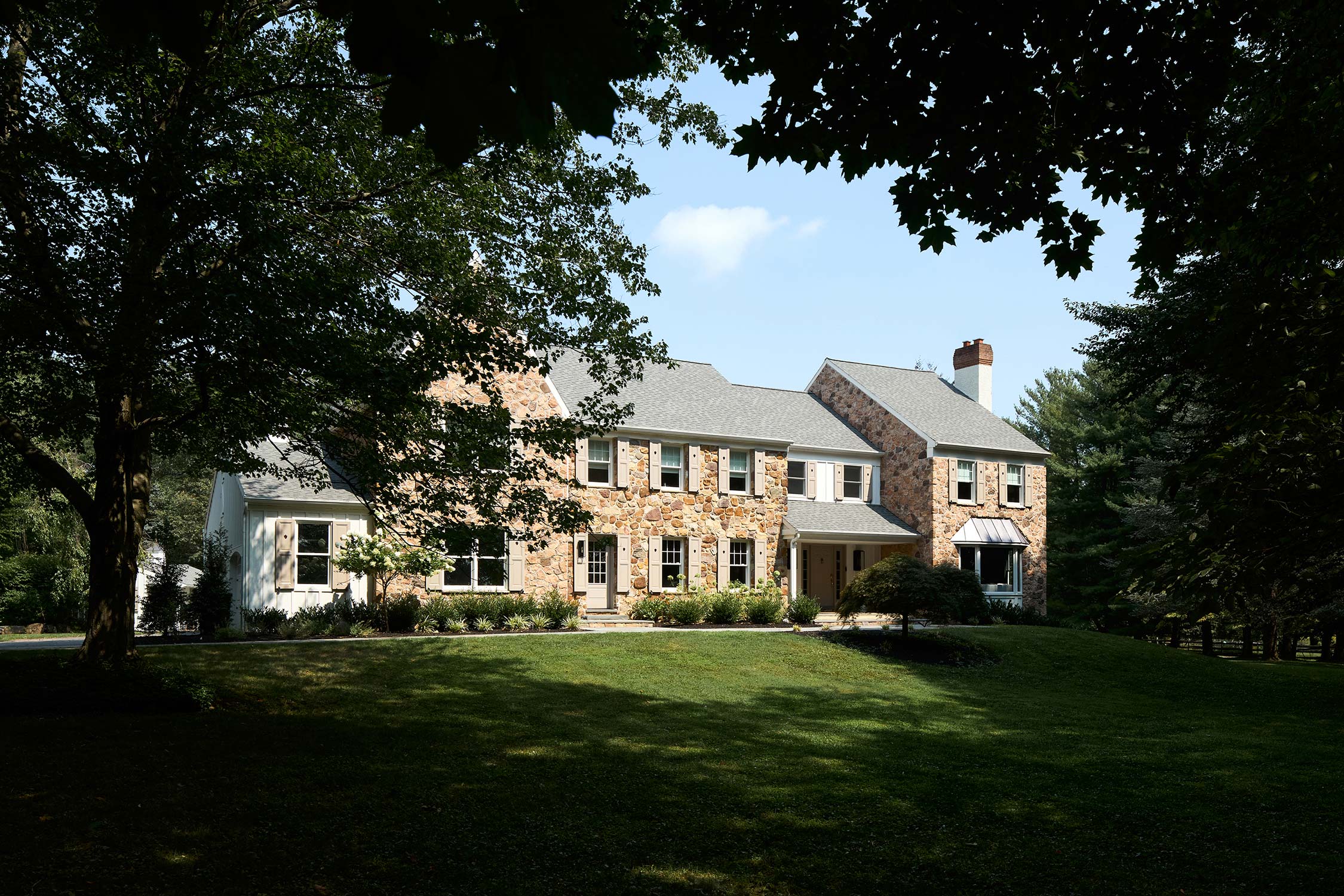Stone house with gray roof, white trim, front porch, and lush green lawn under a partly cloudy sky.
