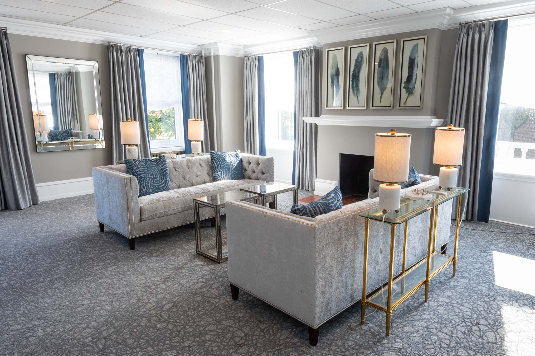 A sophisticated room with gray sofas, blue pillows, and a fireplace. Glass tables, lamps, and framed feather art are set against blue curtains and a patterned rug.