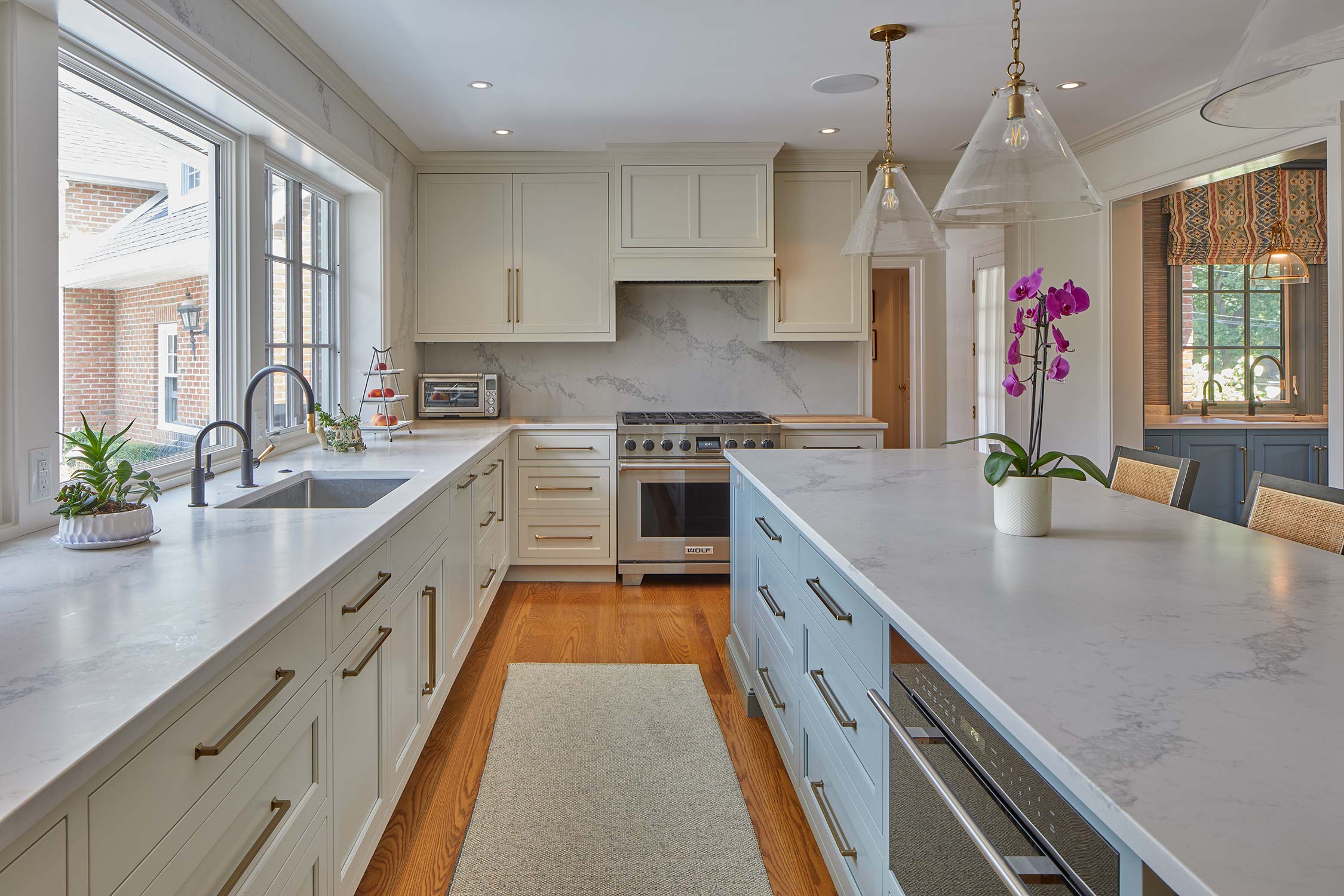 A bright kitchen with white cabinets, a marble island, and pendant lights. A stove, sink, and purple orchids are on the counter, with a window showing a brick house outside.