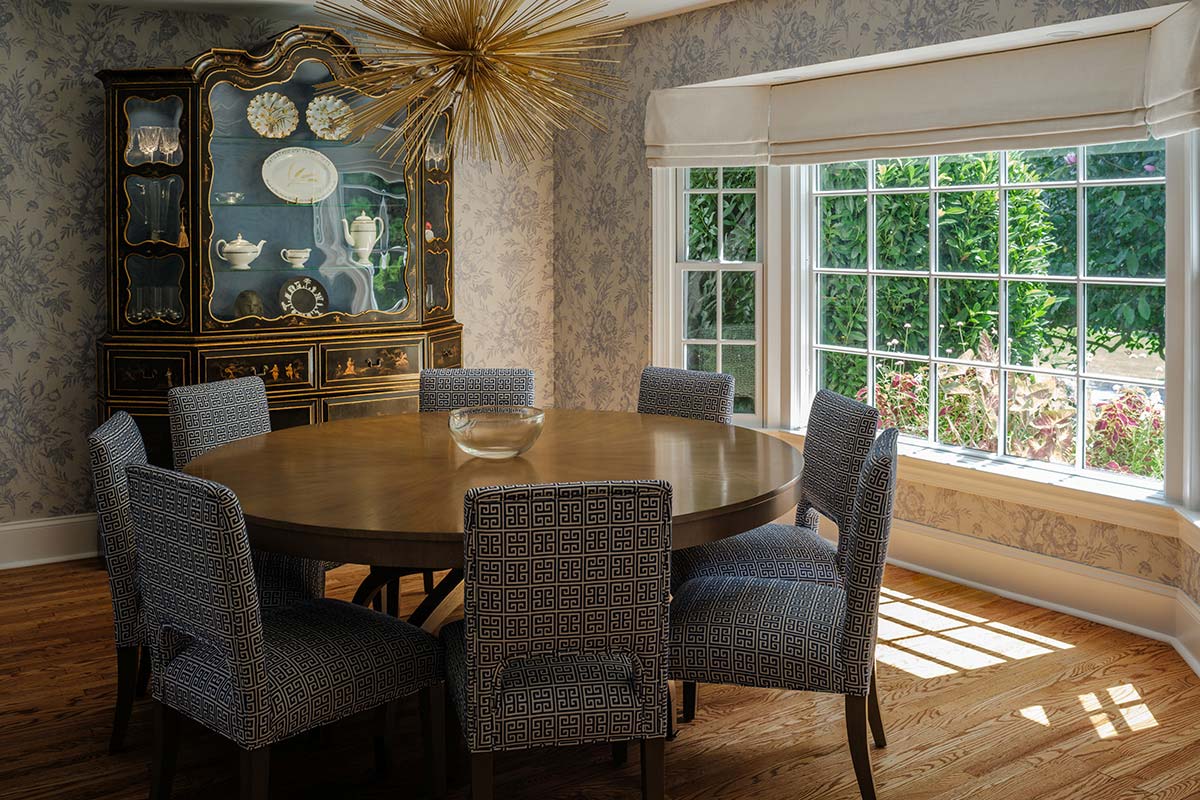 Dining room with a round wooden table, six patterned chairs, and a gold chandelier. A black cabinet with china and large windows with floral wallpaper.
