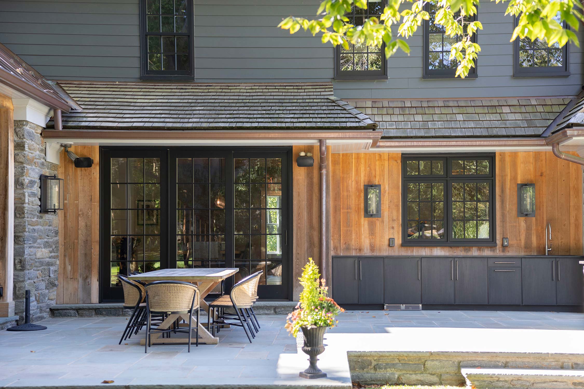 Outdoor patio with a wooden table, wicker chairs, and a potted plant, next to a house with gray siding, large glass doors, and a wooden wall.