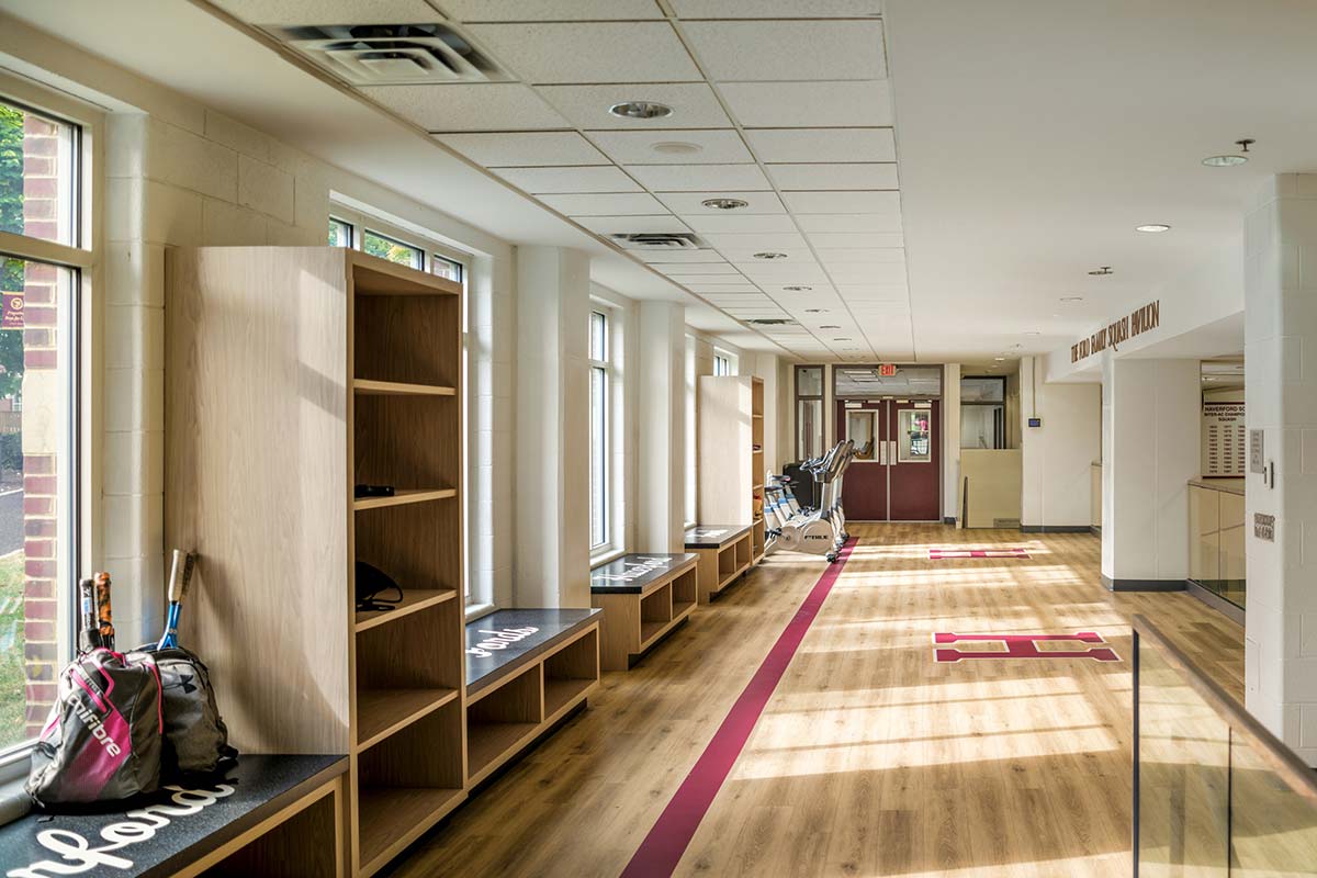 A sunlit hallway with wooden benches, shelves, a backpack, tennis rackets, and an exercise bike near a red door.