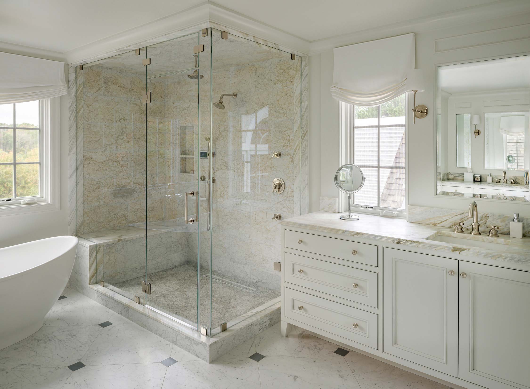 Luxurious bathroom with glass shower, marble walls, freestanding tub, white vanity, and large windows with a scenic view.