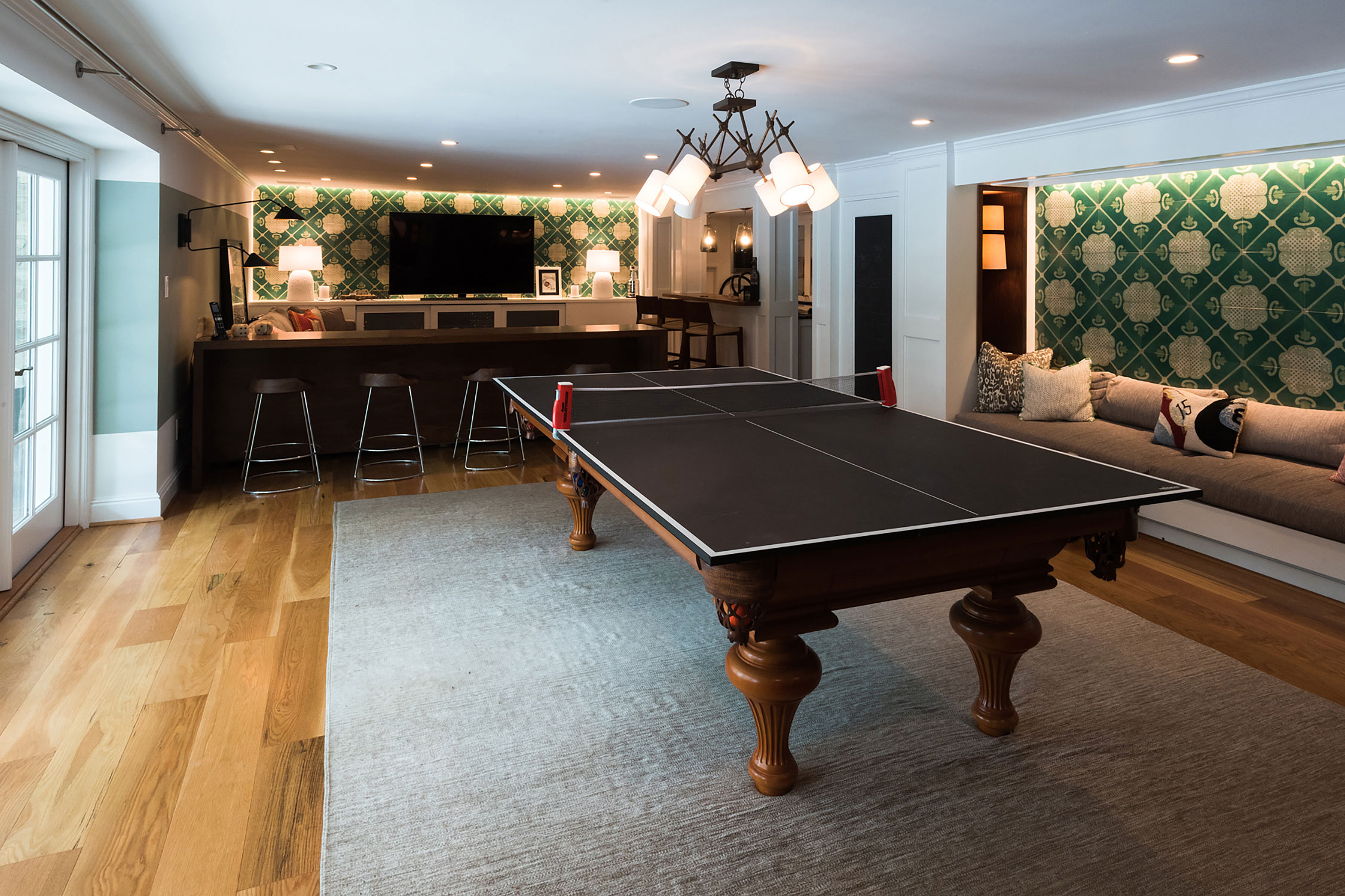 Stylish game room with a sleek ping pong table, modern bar, and cozy seating. Green patterned walls and warm lighting create a vibrant, inviting space.