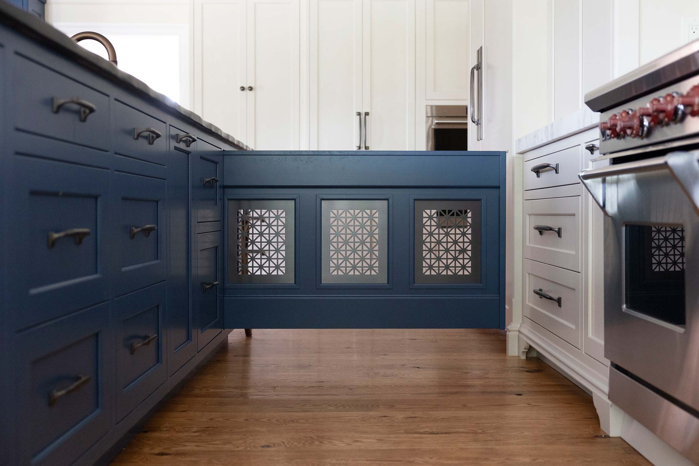 Modern kitchen with blue cabinets, white countertops, and a stainless steel oven. Features intricate lattice panels on a central island over wooden flooring.