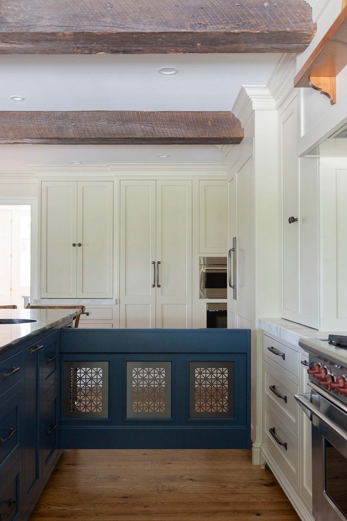 Modern kitchen with a blue island featuring decorative panels, white cabinetry, wooden beams, and a marble countertop on hardwood floors.