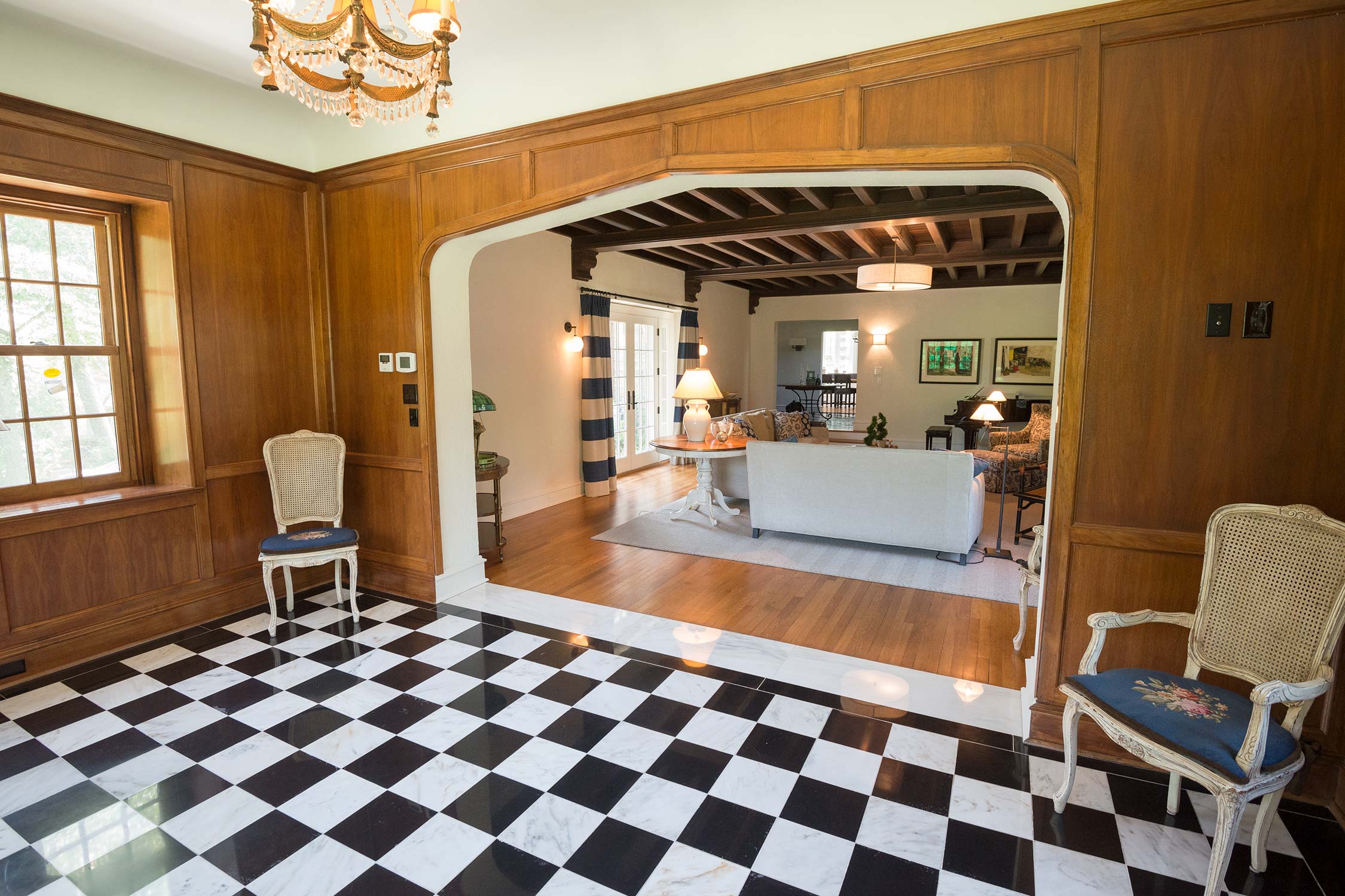 Elegant foyer with black-and-white checkered floor, wooden paneling, chandelier, and view into a stylish living room.