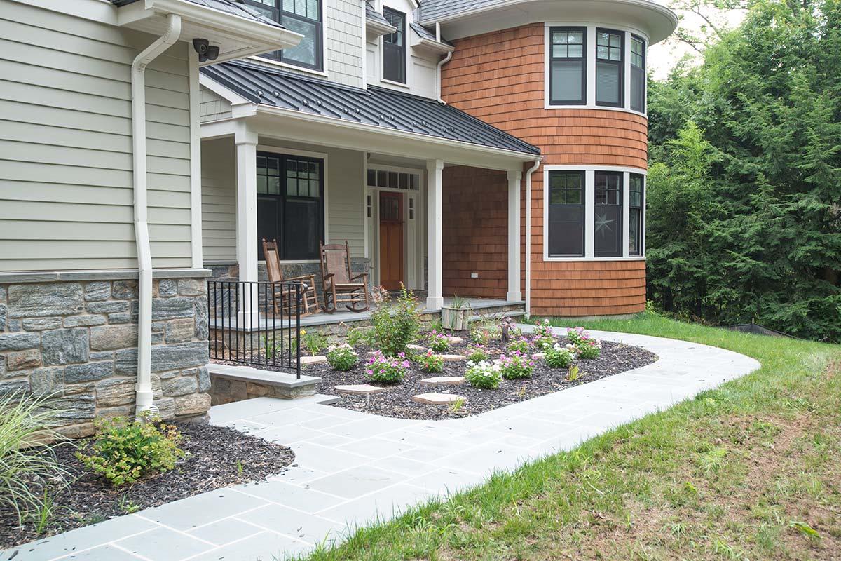 Modern home with a curved orange facade, featuring a stone walkway and vibrant flower garden under a shaded porch.