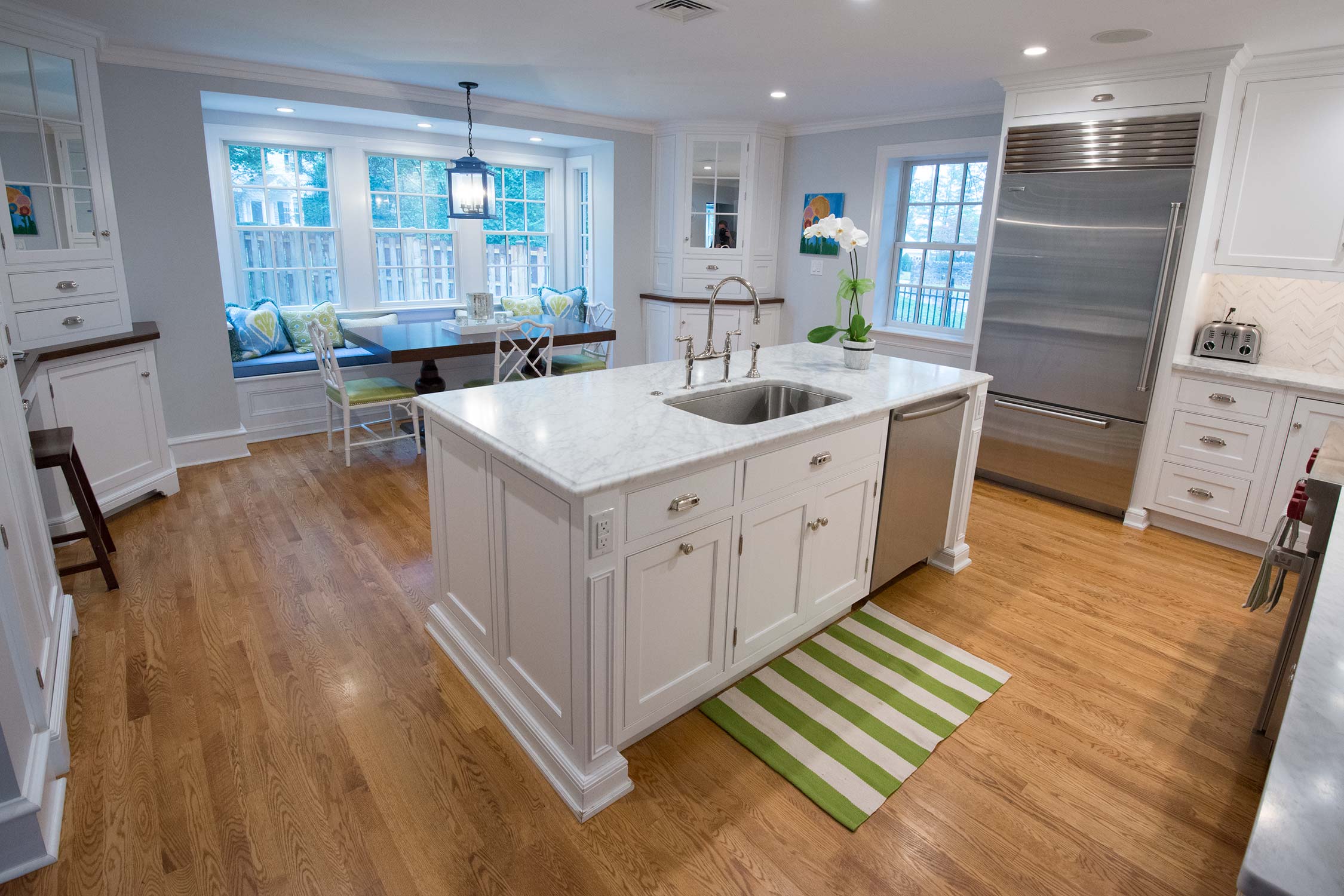 Bright modern kitchen with white cabinets, marble island, stainless steel appliances, and large windows. Features a cozy dining nook with colorful cushions.