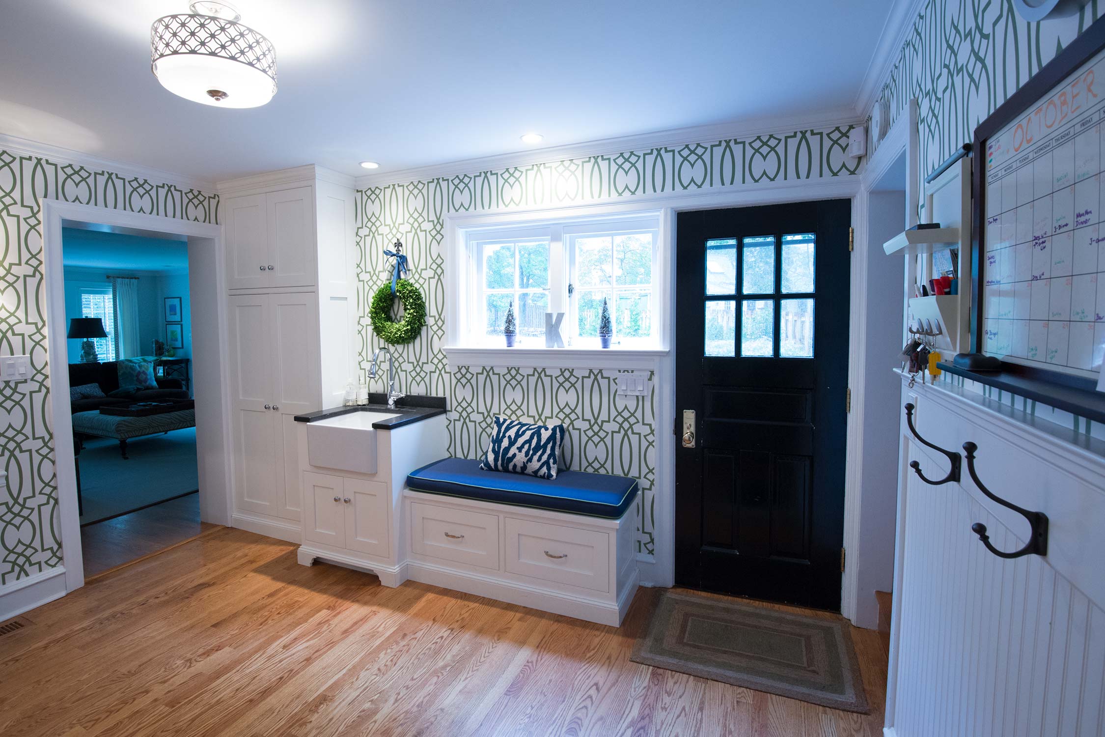Stylish entryway with white cabinet, blue bench, and patterned wallpaper. Features a black door, window decor, and a wall calendar for organization.