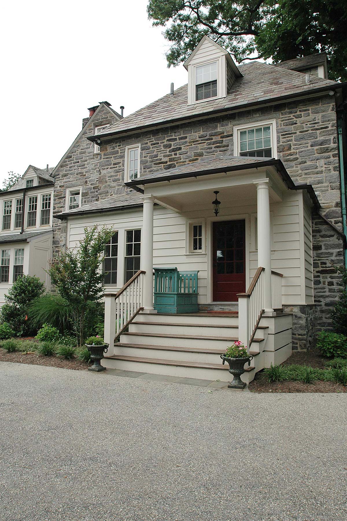 Charming stone house with a white porch, red door, and teal bench. Surrounded by lush greenery and potted plants, offering a welcoming entrance.