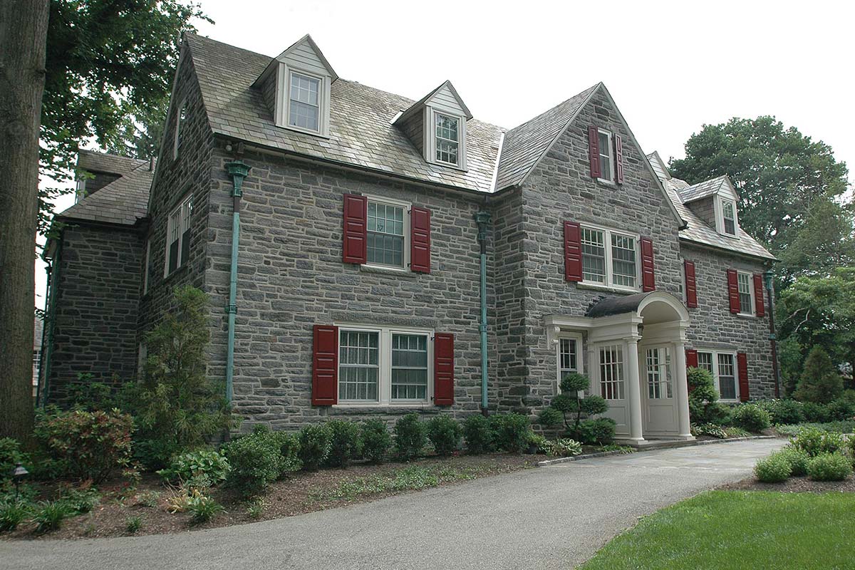 Elegant stone mansion with red shutters and a white entrance. Surrounded by lush greenery and a curved driveway, exuding timeless charm.