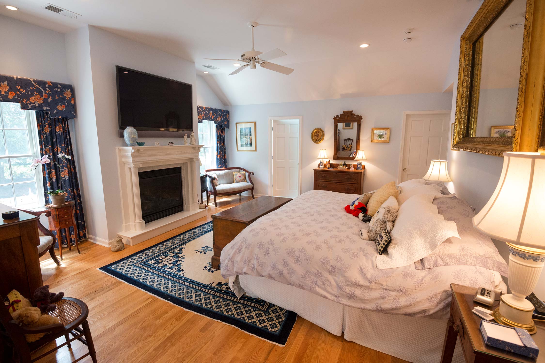 Spacious bedroom with king bed, fireplace, mounted TV, and ceiling fan. Features hardwood floors, floral curtains, antique furniture, and cozy decor.