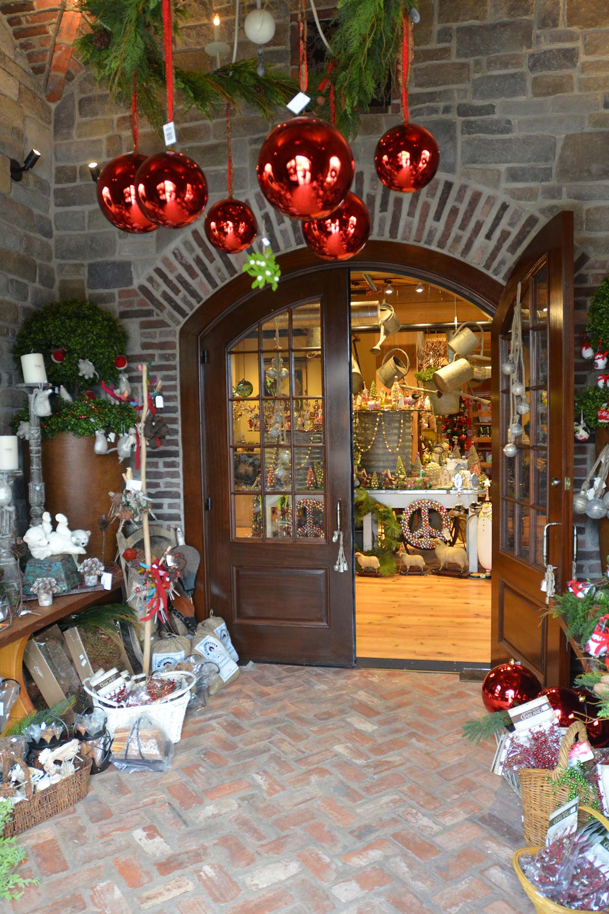 Festive stone entrance with wooden doors, garlands, red ornaments, wreaths, and holiday decor items on a brick floor.