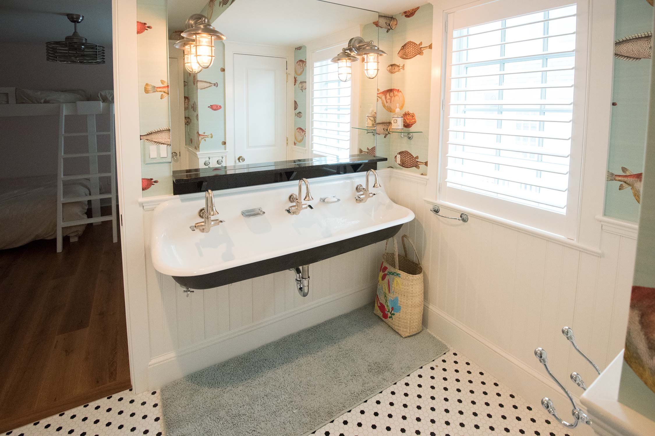 A bathroom with a double sink, fish-themed wallpaper, two wall sconces, white shutters, and black-and-white tiled floor.