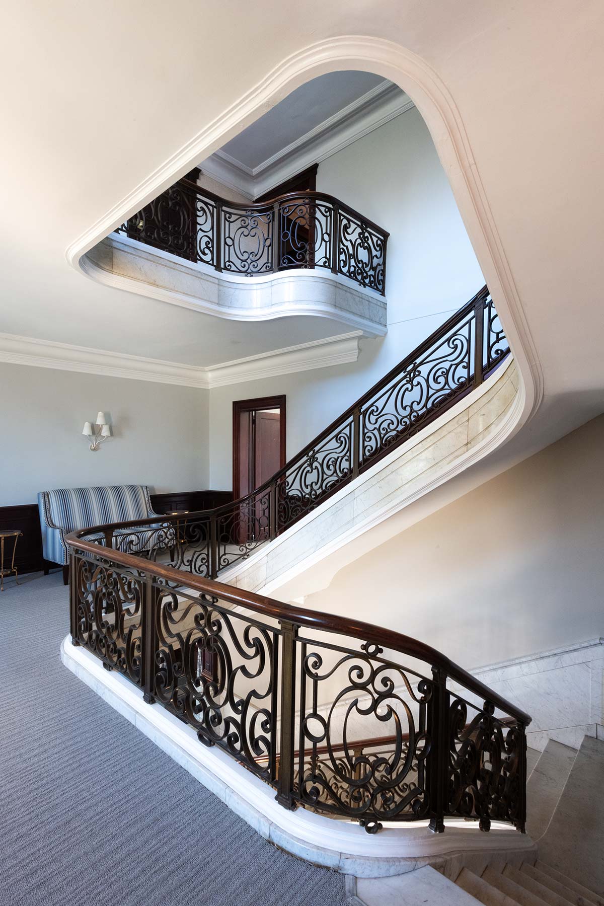 A grand marble staircase with ornate black iron railings winds through a hallway. A striped blue sofa sits near a wooden door, with a wall sconce above.
