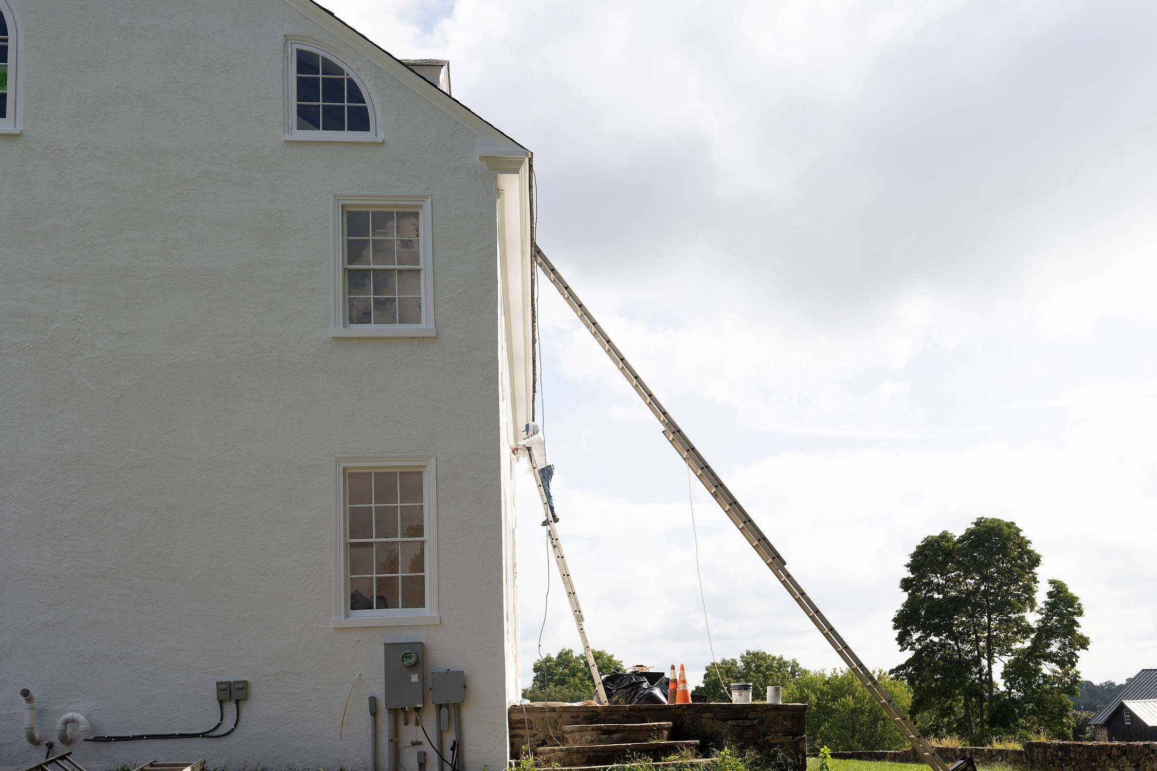 Worker on ladder painting a white house with arched window, near construction materials, trees, and a stone wall under a cloudy sky.
