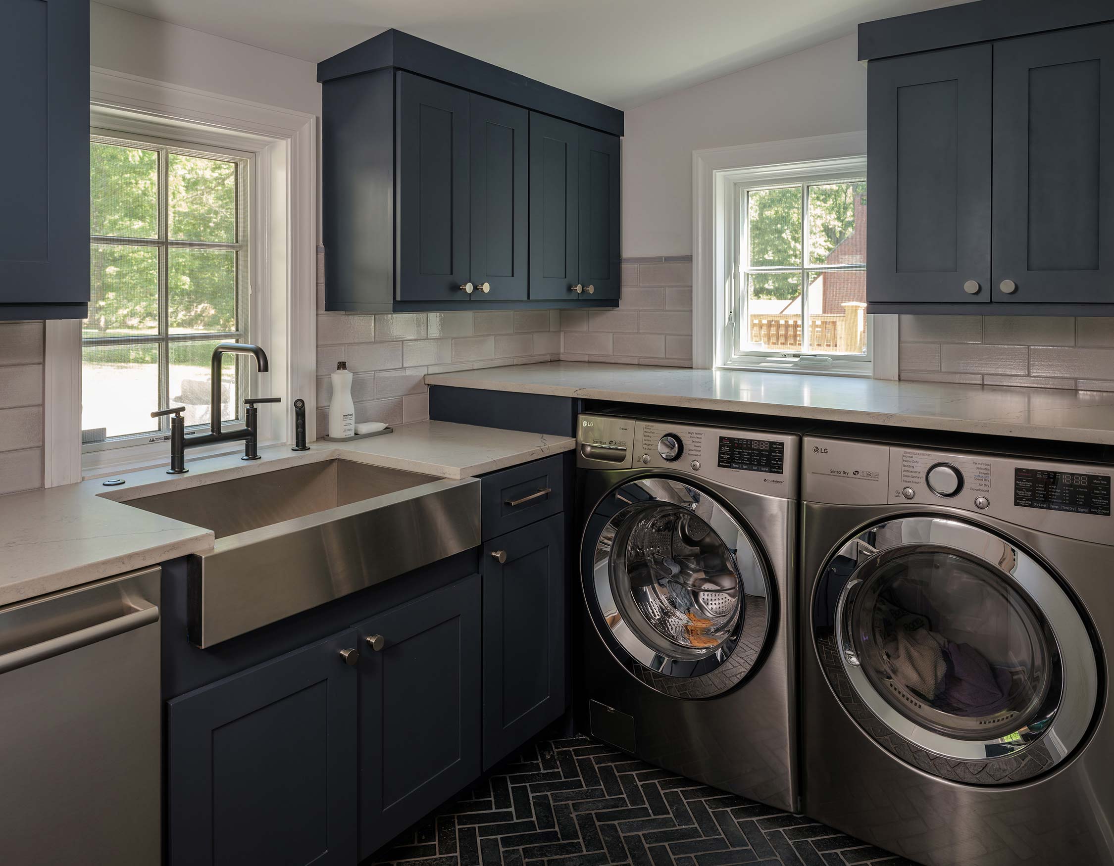 Laundry room with dark blue cabinets, a stainless steel sink, and two front-load washers. Subway tile backsplash and large windows let in natural light.