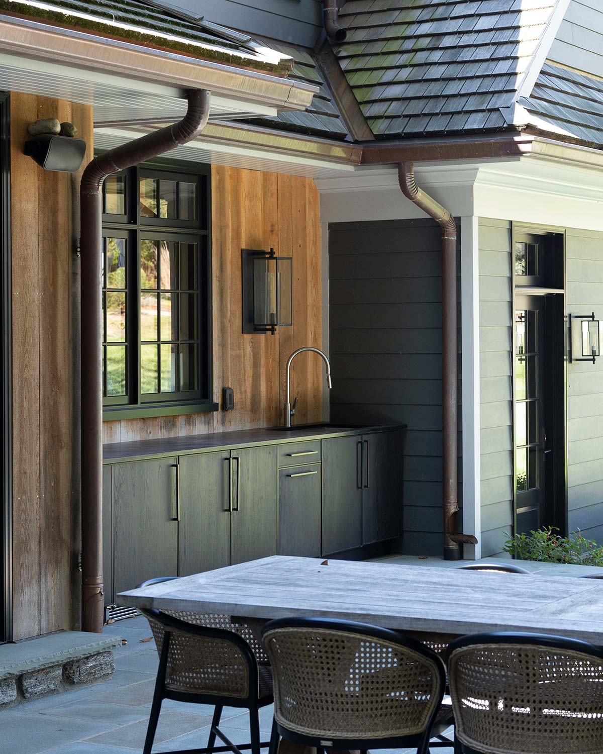 Outdoor kitchen with dark cabinets, sink, and a wooden table with wicker chairs, next to a house with shingled roof and wooden walls.