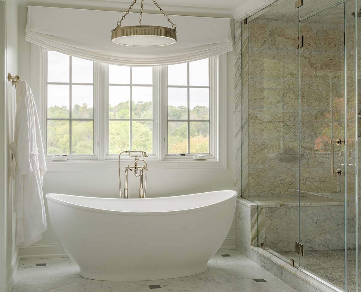 Elegant bathroom with a freestanding white tub, glass shower, marble walls, large windows with greenery view, and hanging light.