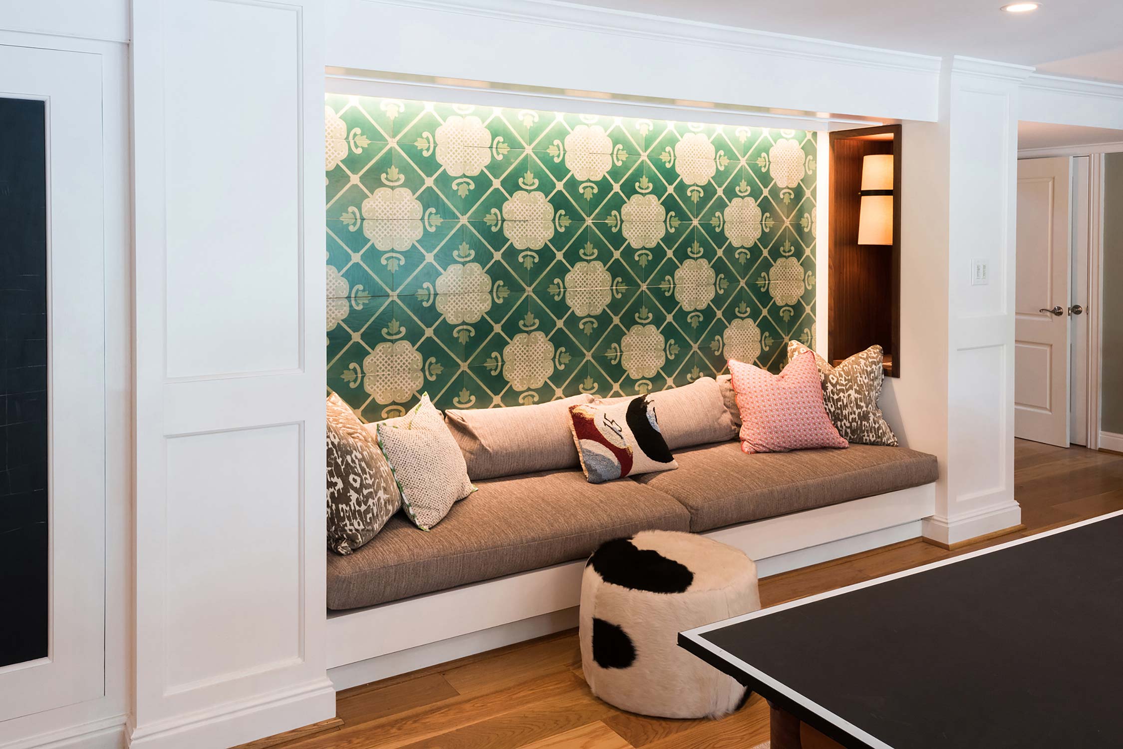 Cozy nook with a cushioned bench, vibrant green patterned wall, and colorful pillows. Includes a unique cowhide ottoman and a sleek ping pong table.