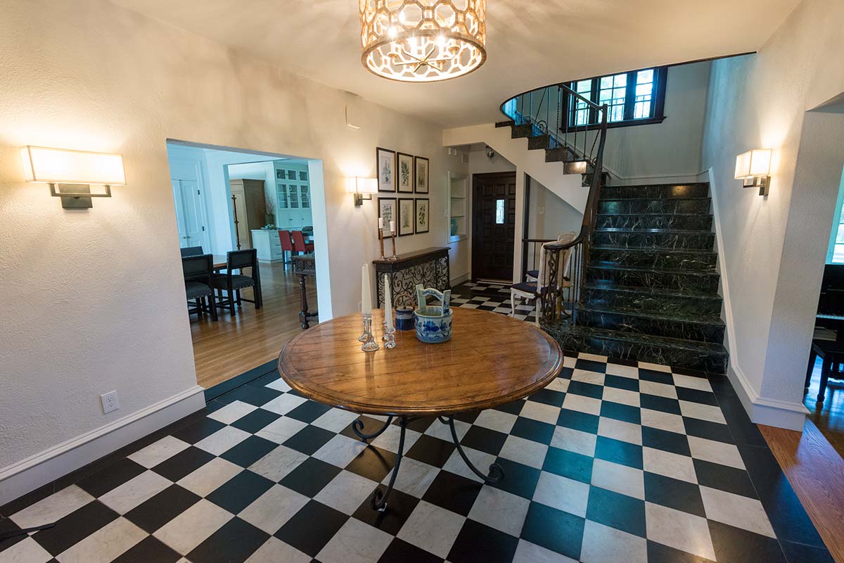 Grand foyer with black-and-white checkered floor, wooden table, chandelier, and elegant staircase with marble steps.