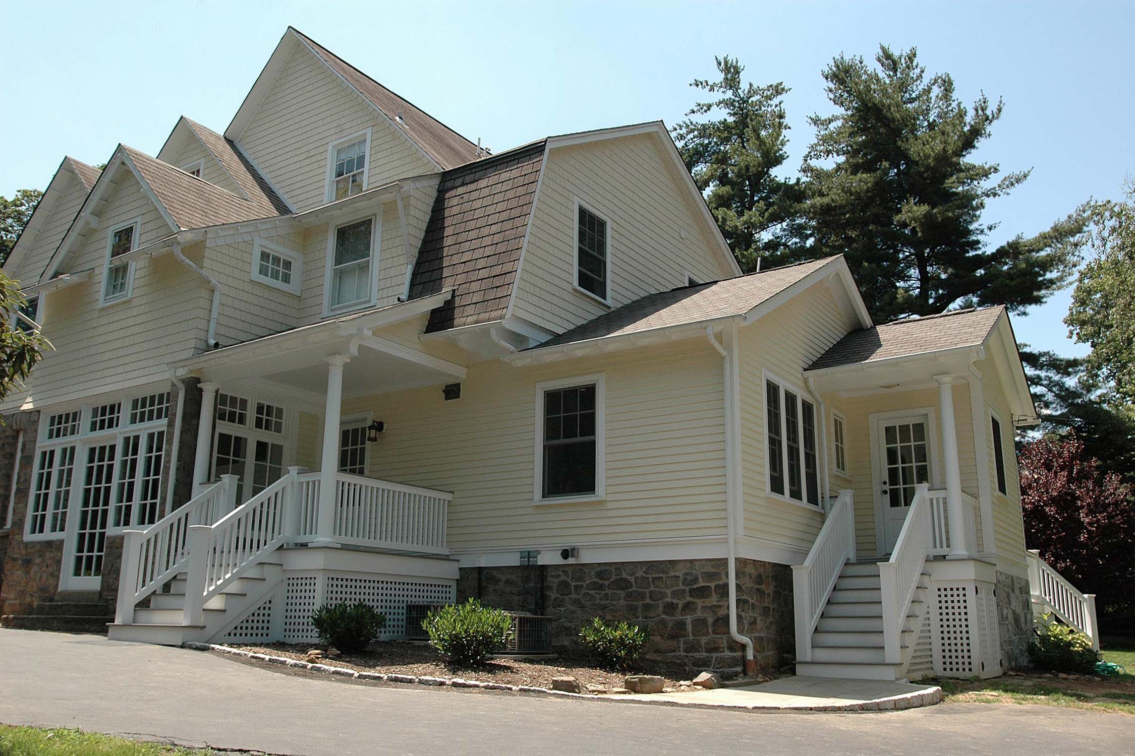 Charming pale yellow house with white porches and stone base. Surrounded by lush trees and shrubs, featuring multiple gabled roofs.