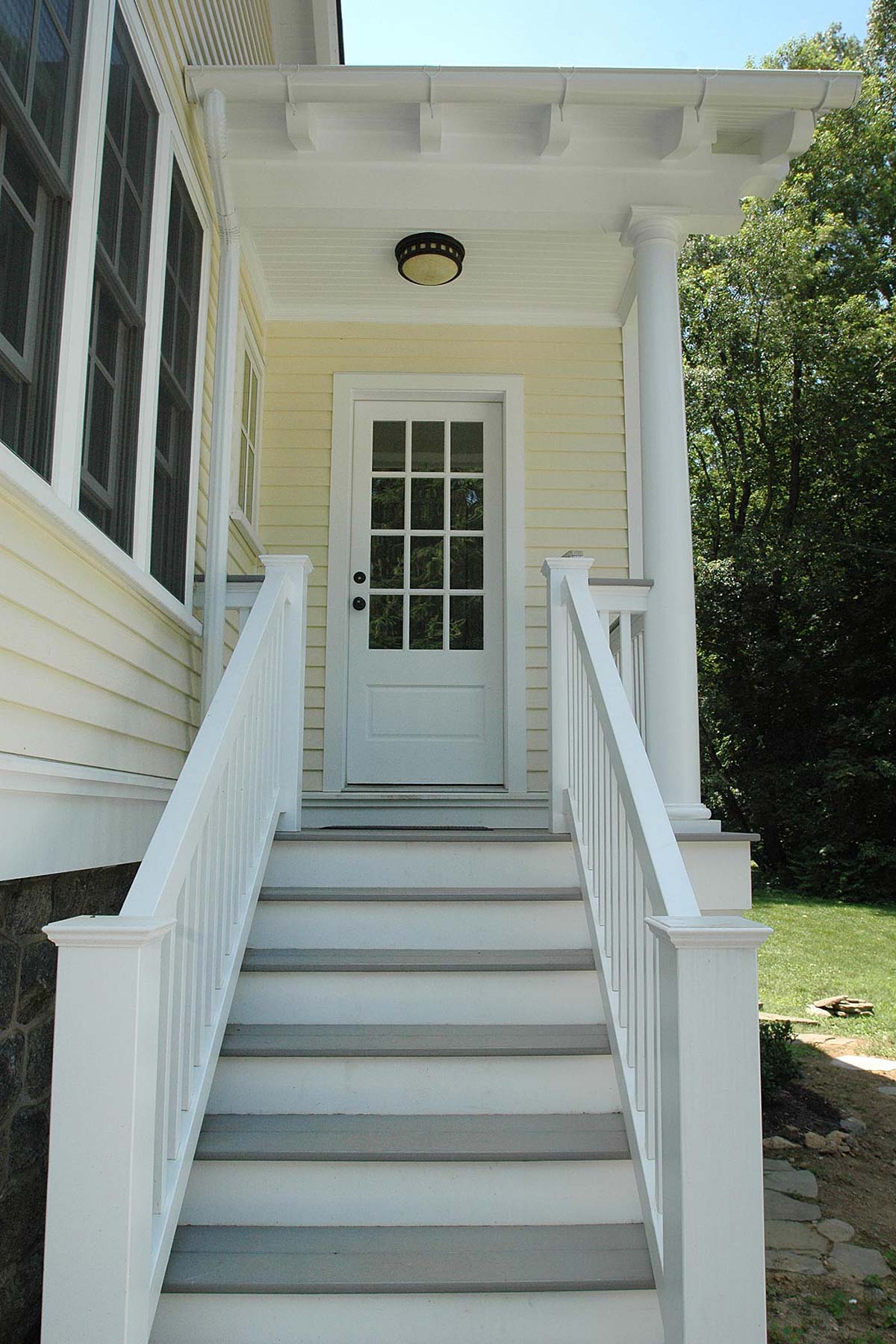 Inviting pale yellow home with a white porch and staircase. Features a glass-paneled door and lush greenery, creating a warm entrance.