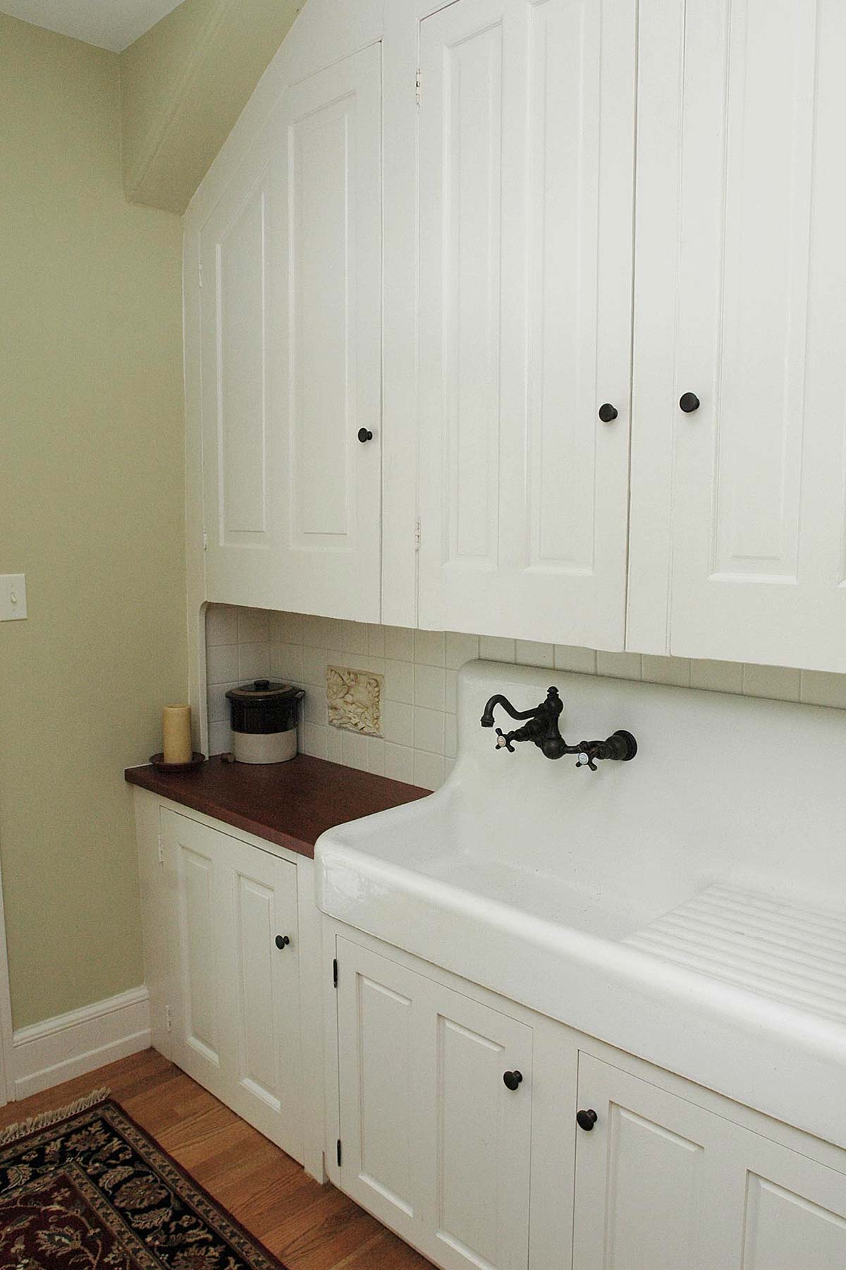 Cozy kitchen nook with white cabinets and a large white sink. Features a dark faucet, tiled backsplash, and warm wood countertop.