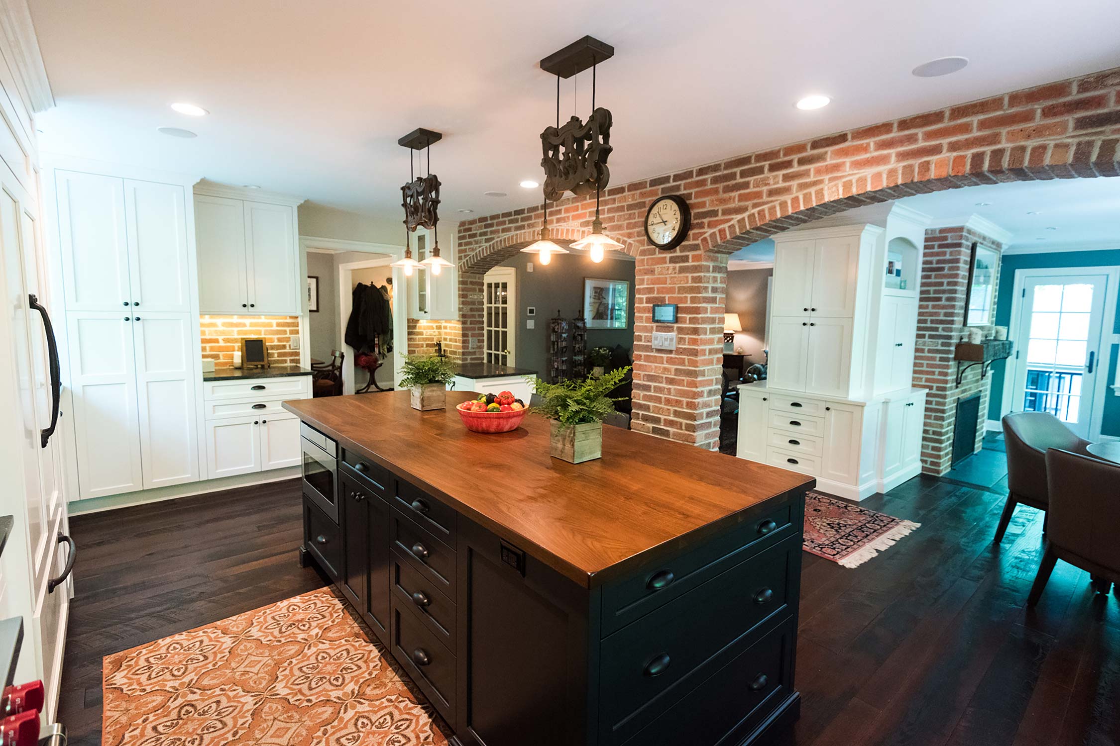 Modern kitchen with wooden island, brick archway, white cabinets, and rustic decor, featuring a clock and cozy dining area.