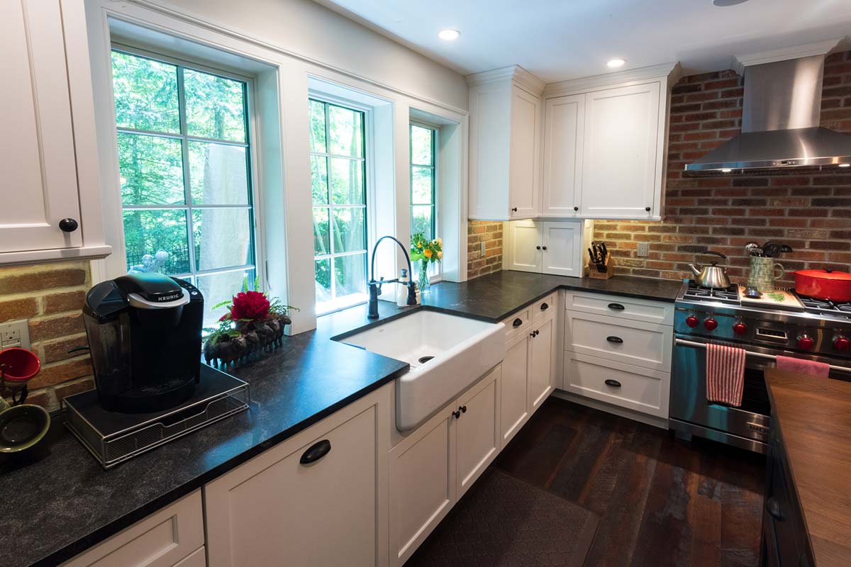 Bright kitchen with white cabinets, black countertops, brick backsplash, and large windows, featuring a farmhouse sink and modern appliances.