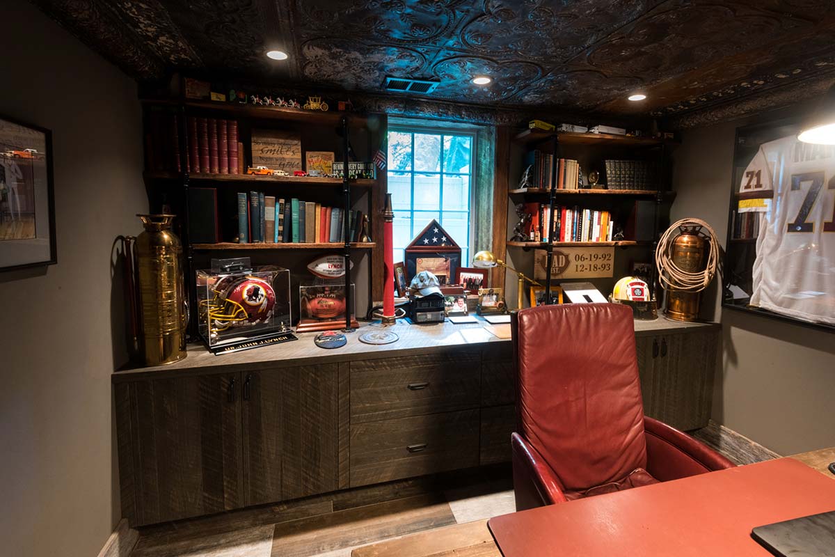 Cozy office with rustic wooden shelves, sports memorabilia, and a red leather chair, featuring a decorative tin ceiling and natural light.