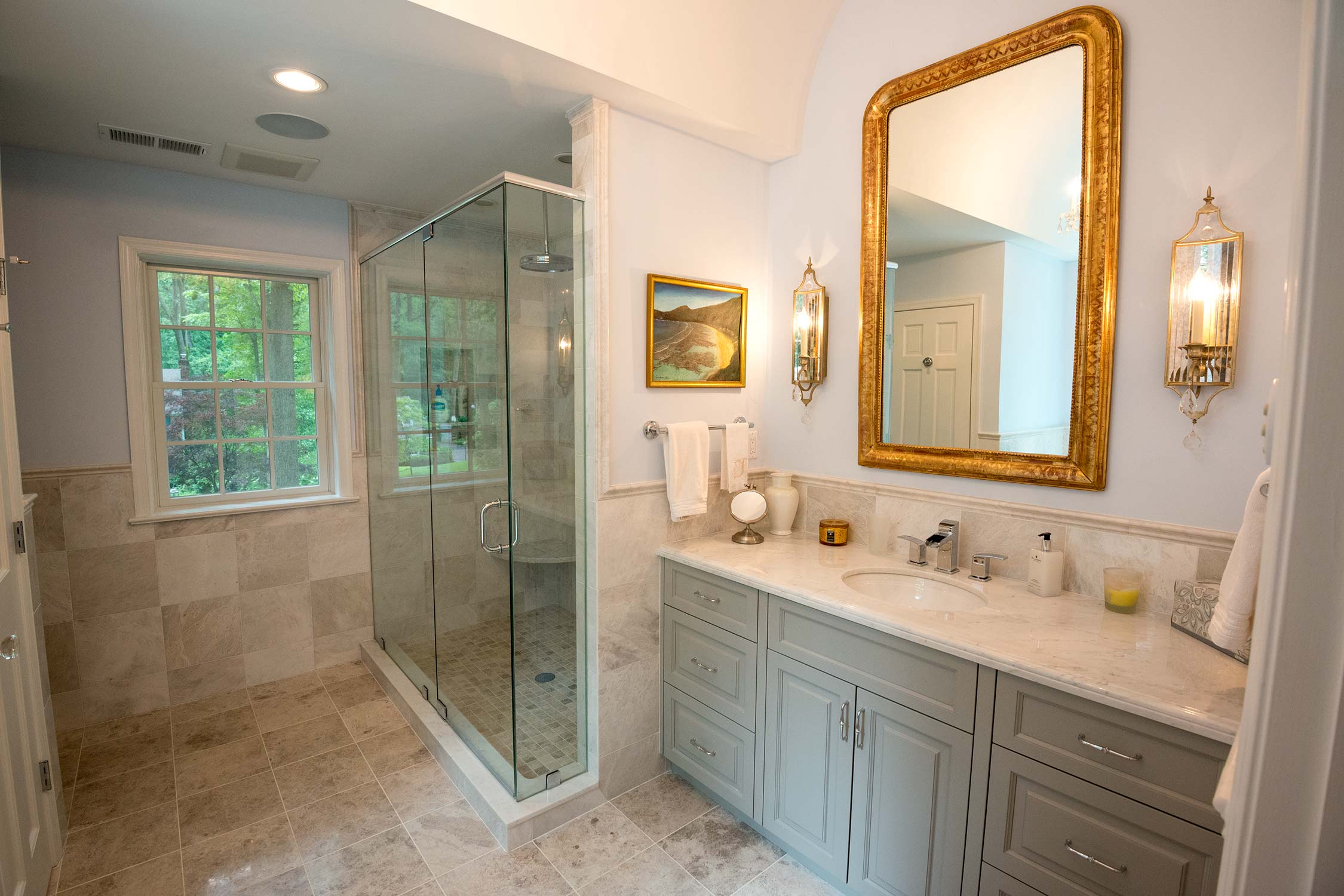 Luxurious bathroom with glass shower, marble vanity, and large gold mirror. Features tiled floors, natural light, and elegant wall sconces.