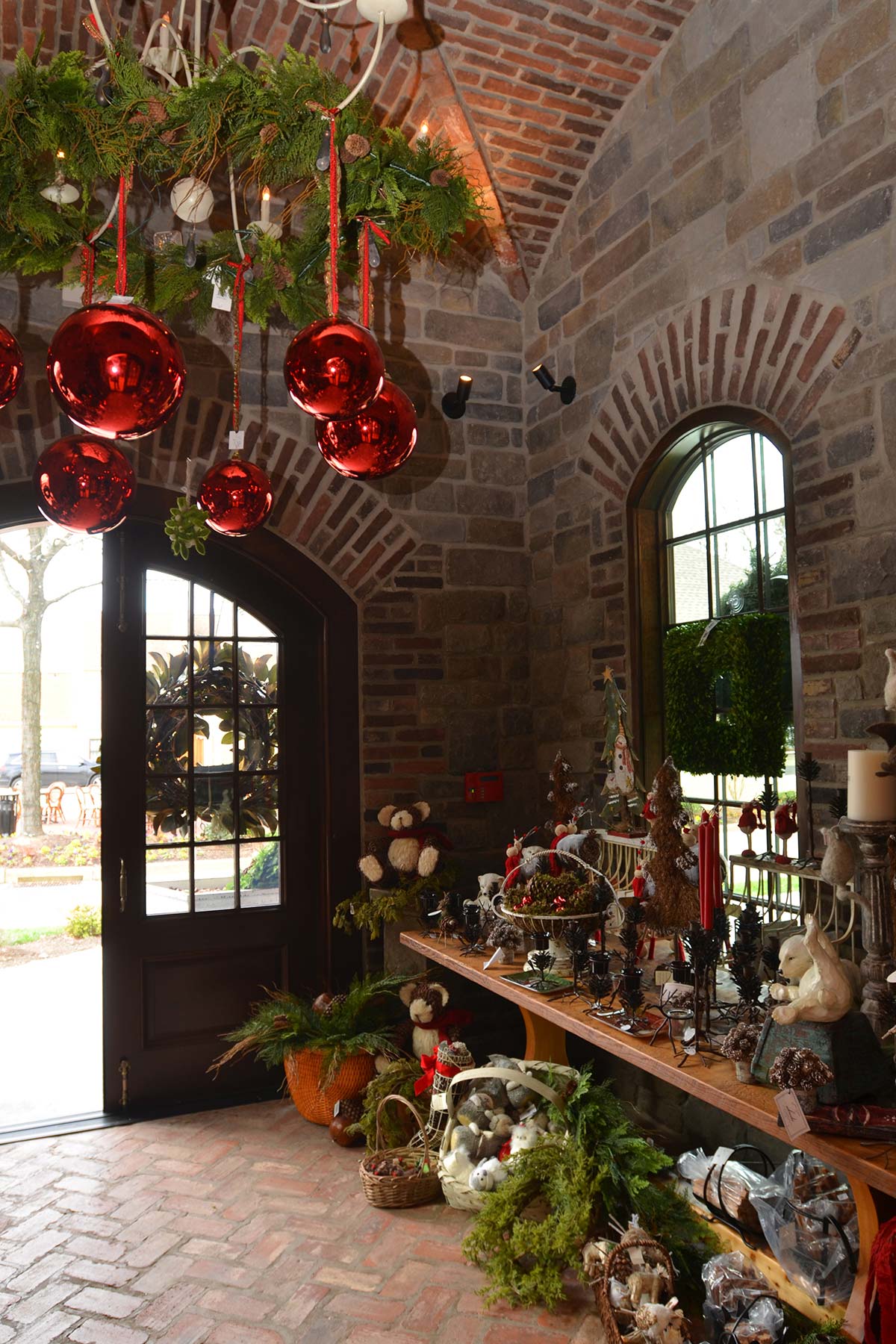 Festive stone entrance with wooden doors, garlands, red ornaments, wreaths, and holiday decor items on a brick floor.