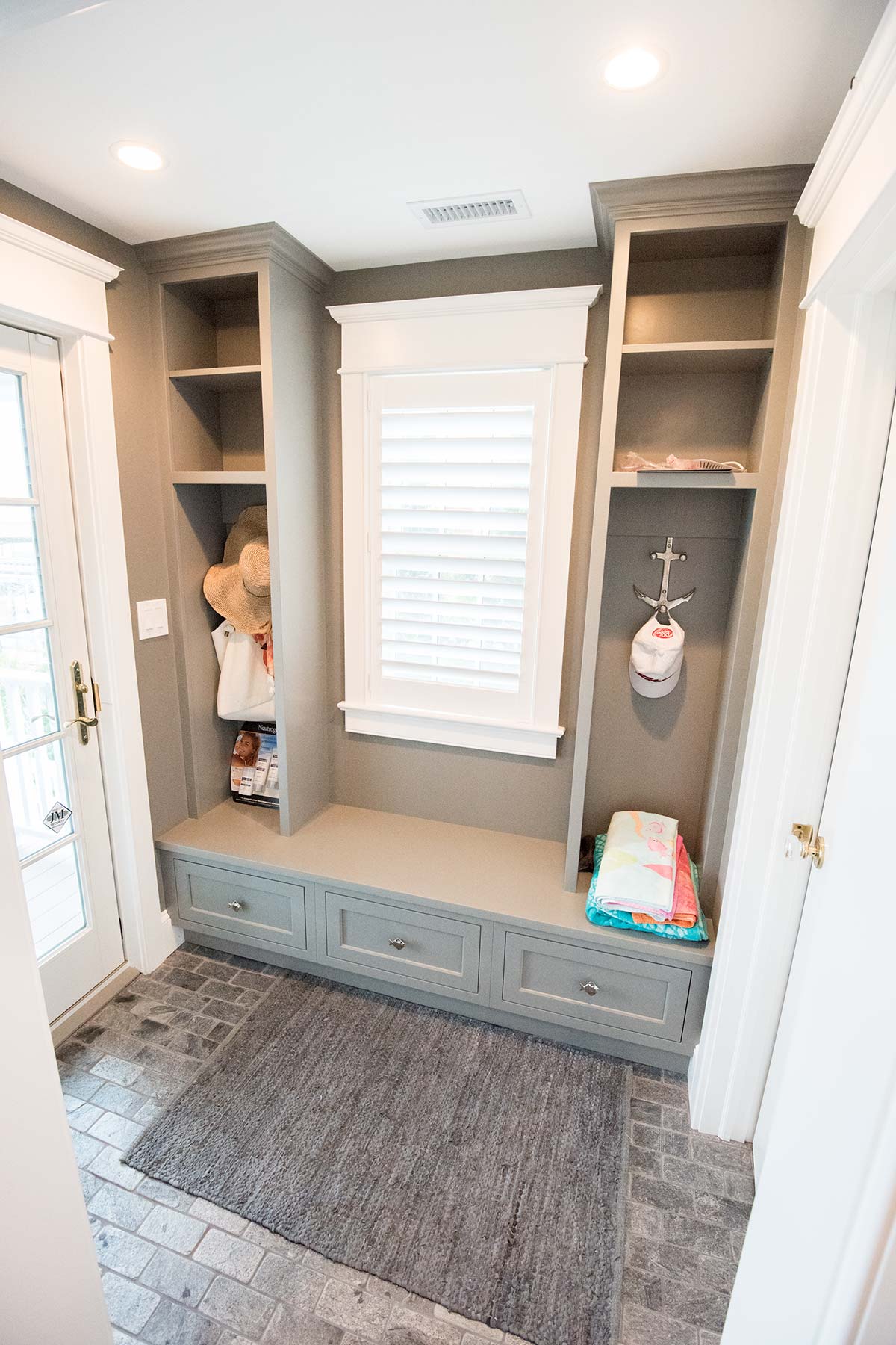 A mudroom with a gray bench, drawers, open shelves, hats, towels, an anchor hook, and stone tile floor.