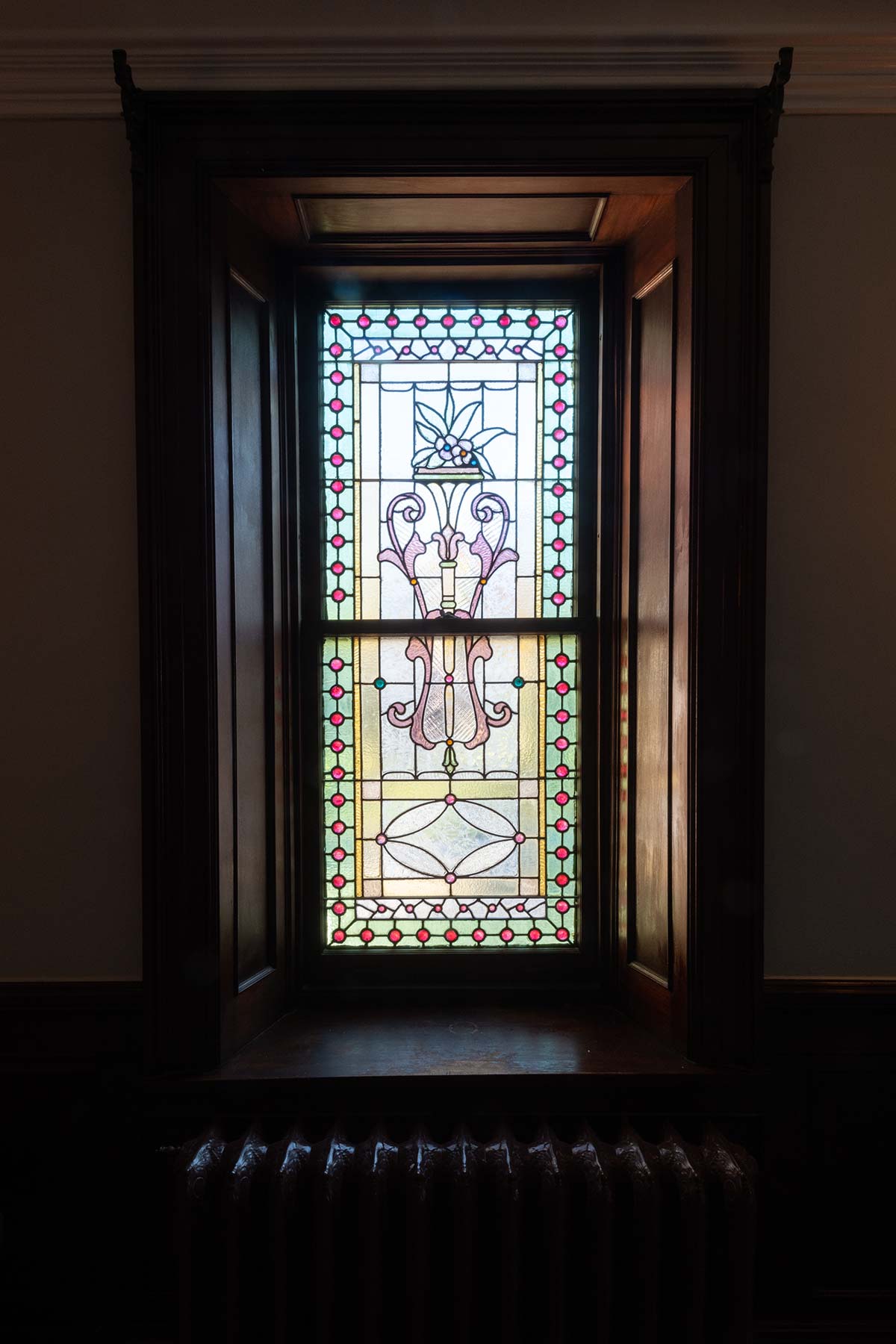 A tall stained glass window with colorful floral designs, set in a wooden frame. A radiator sits below, and sunlight filters through the intricate patterns.