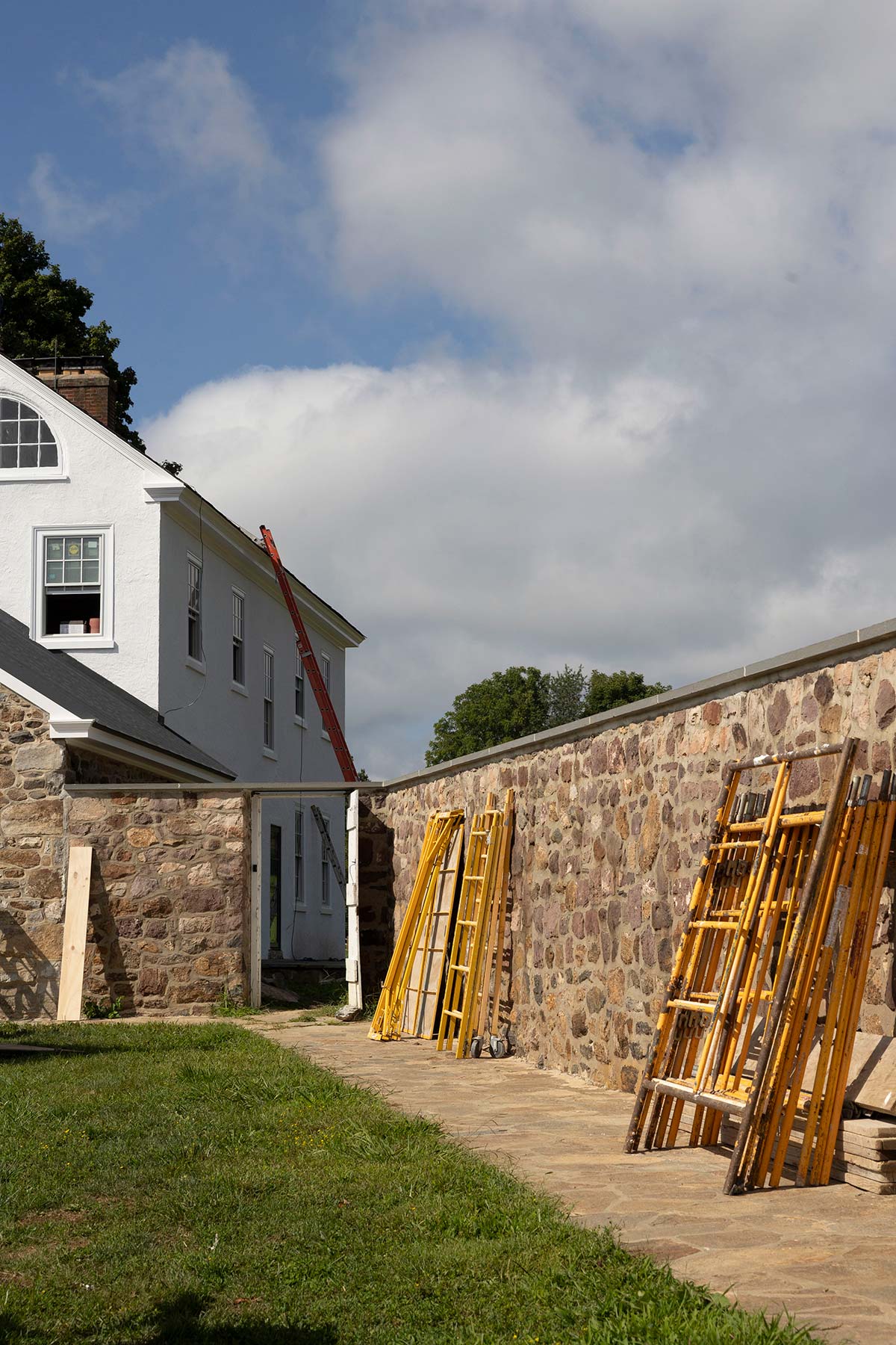 White house with stone wall, ladders, and construction materials, surrounded by grass and trees under a cloudy sky.