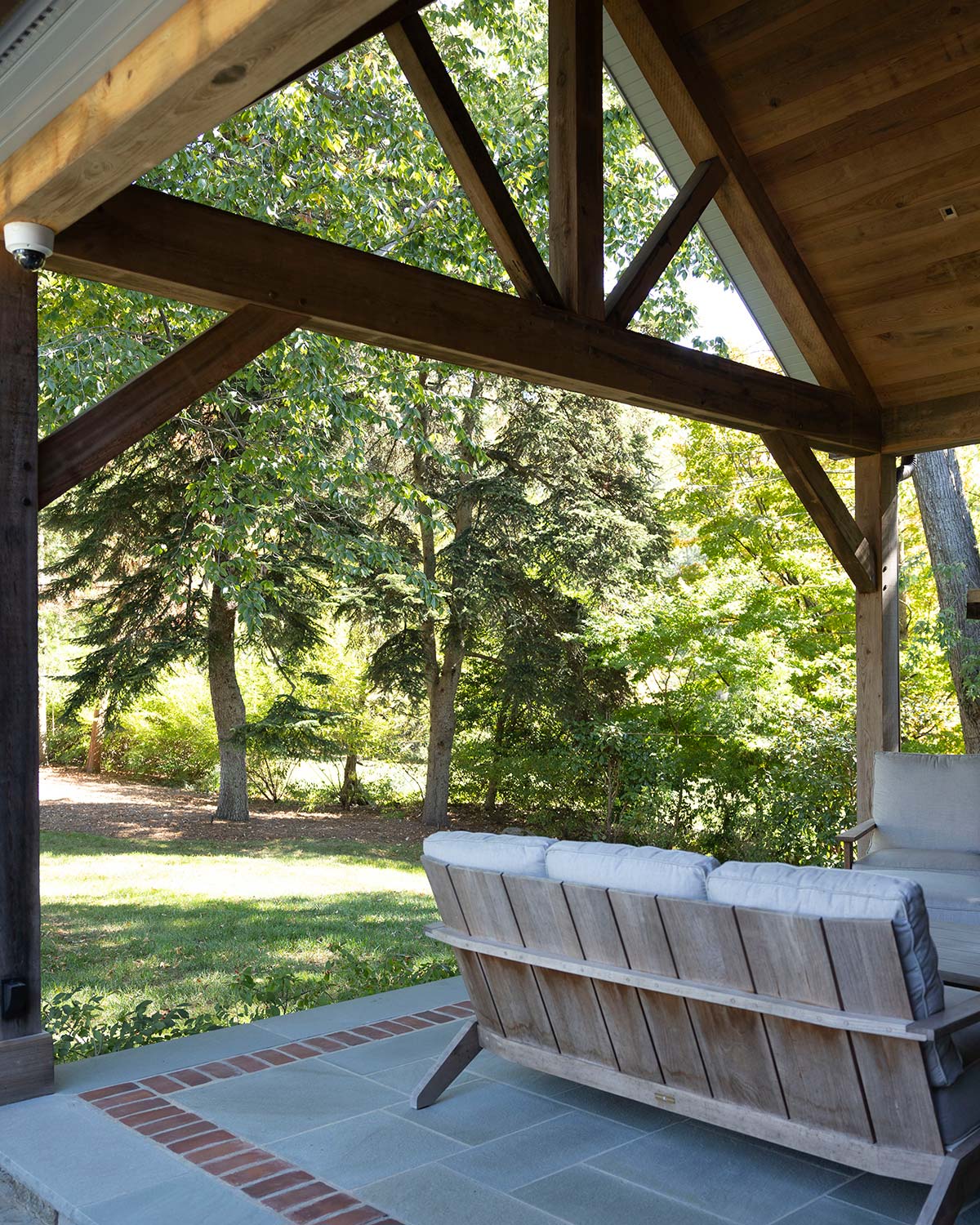 Covered patio with wooden beams and a cushioned wooden bench, overlooking a lush green lawn and tall trees.