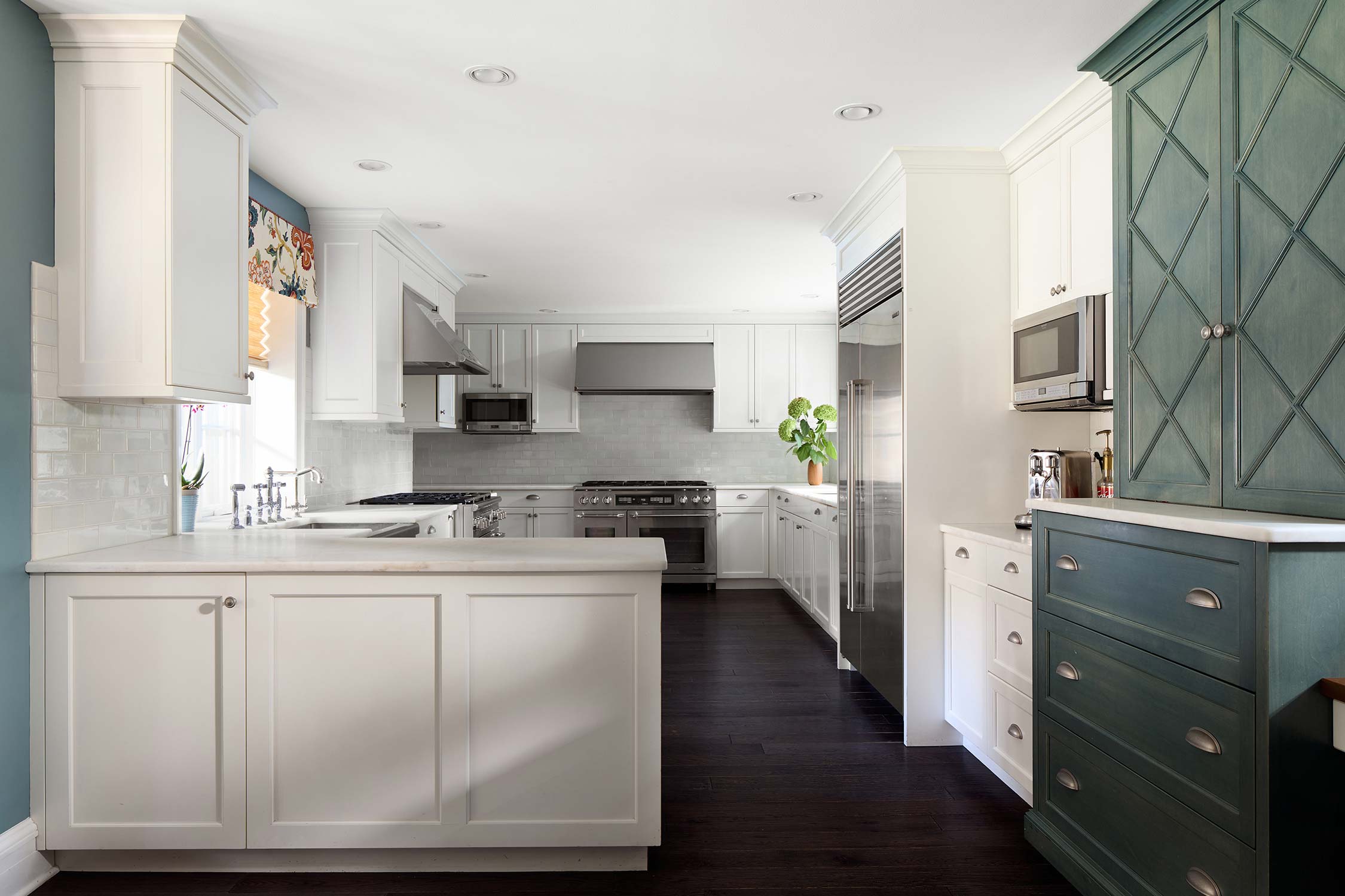 Modern kitchen with white and teal cabinets, stainless appliances, marble counters, subway tile backsplash, and floral shades.