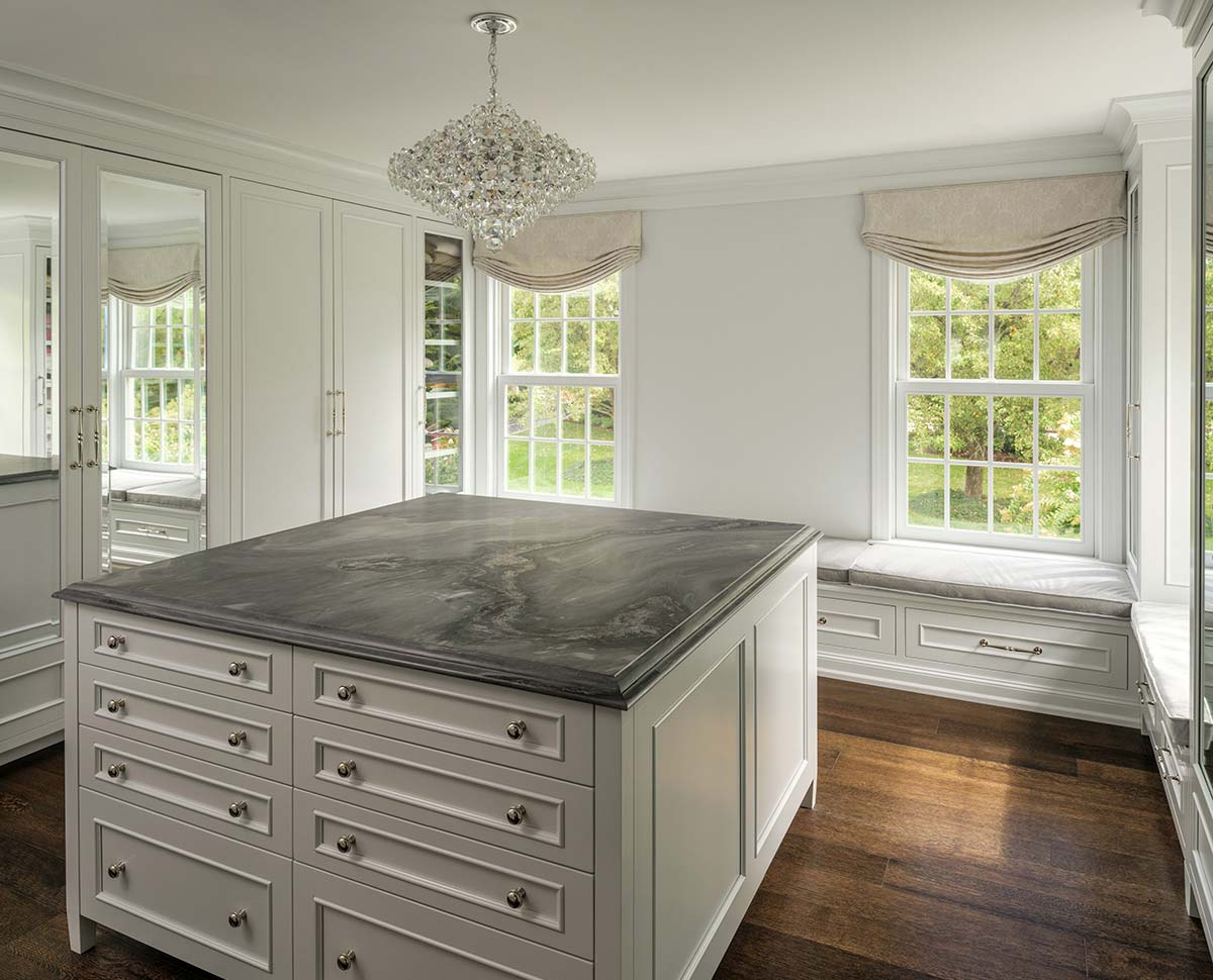 Elegant dressing room with a marble-top island, white cabinets, crystal chandelier, window seat, and large windows with greenery view.