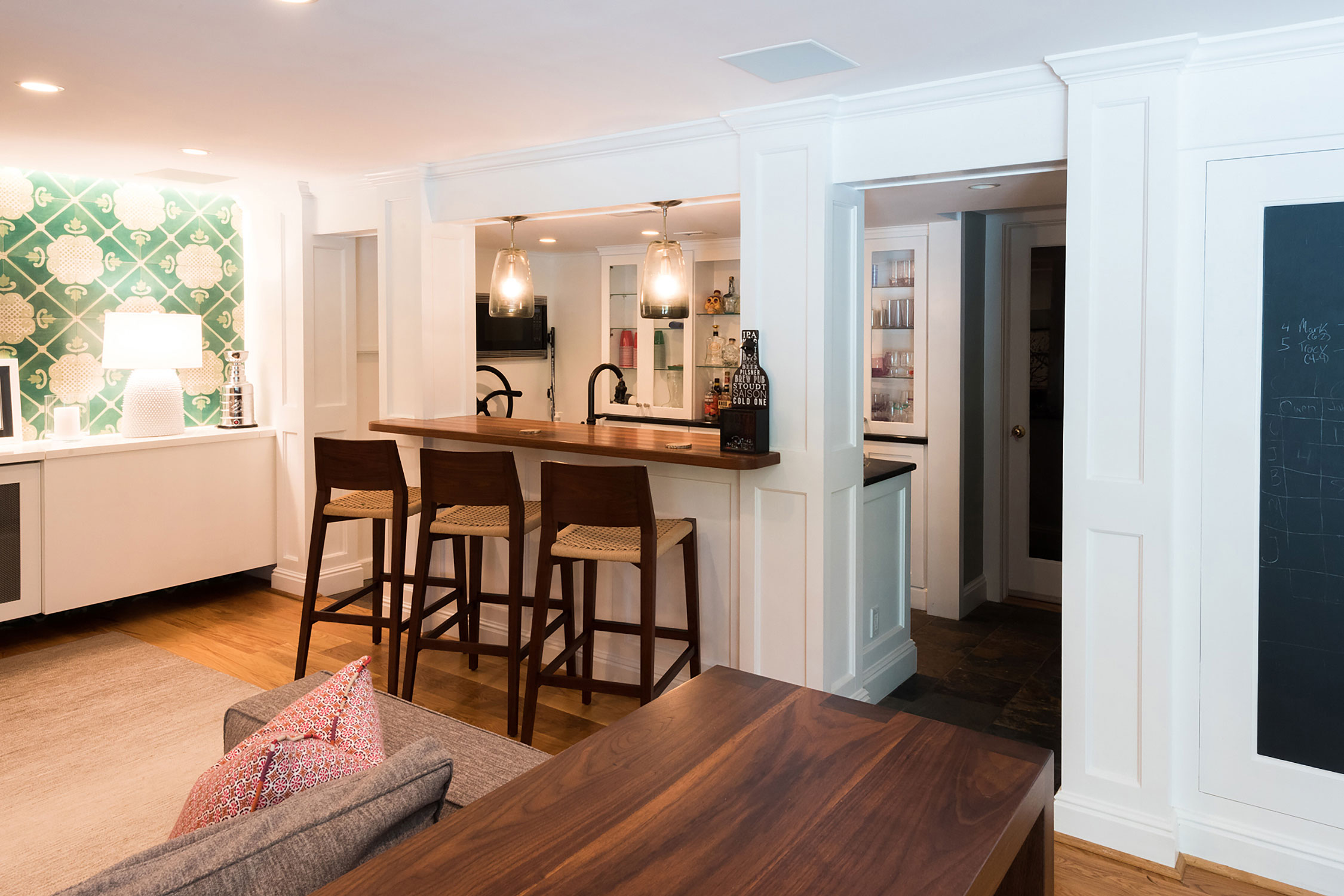 Stylish bar area with a wooden counter, modern stools, and a green patterned wall. Features a cozy sofa, shelves with decor, and warm lighting.