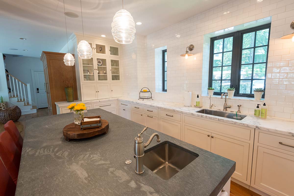 Modern kitchen with white cabinets, gray stone countertops, pendant lights, and large windows overlooking greenery.