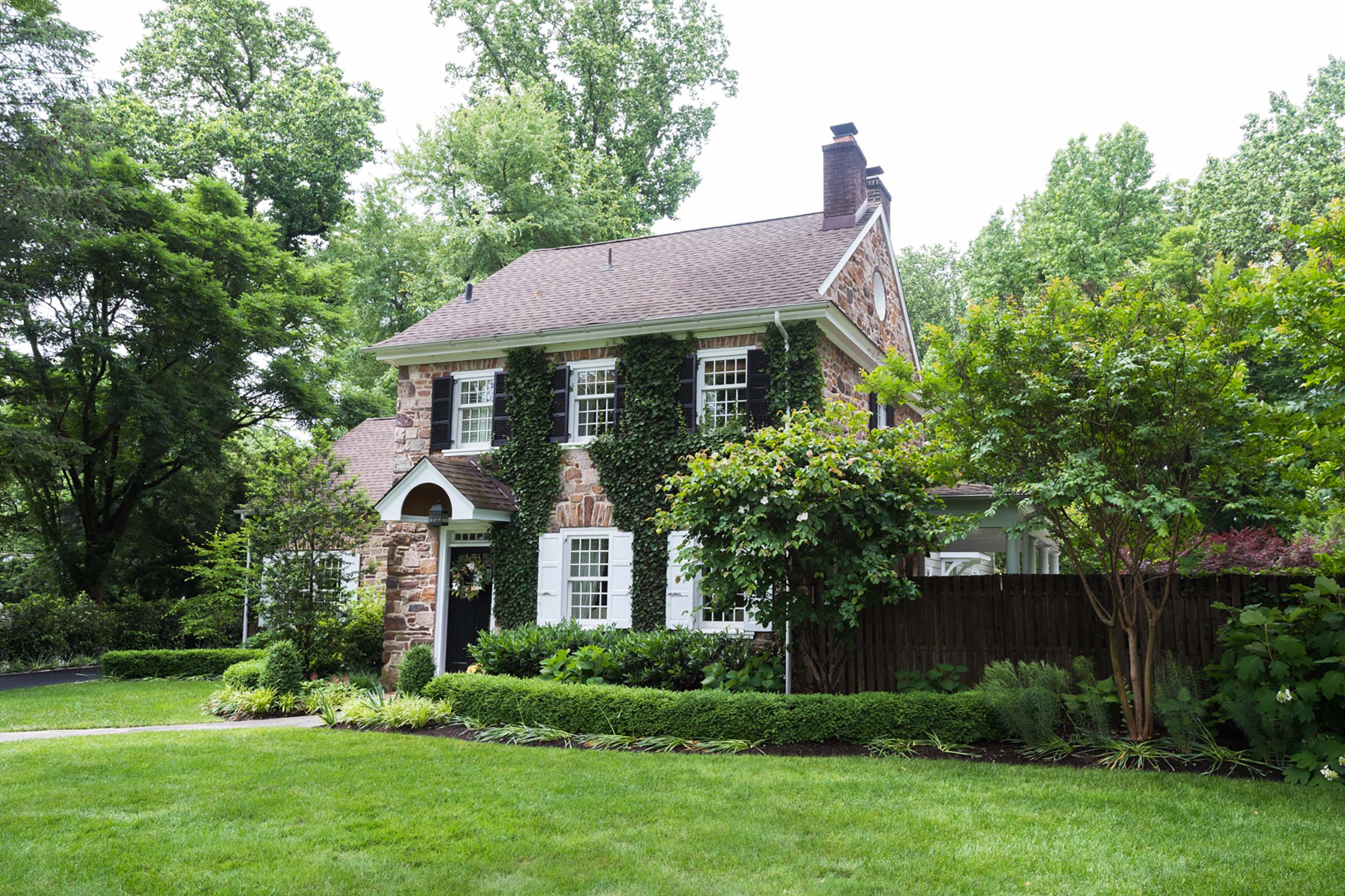 Quaint stone house surrounded by lush greenery and trees, featuring ivy-covered walls, a well-manicured lawn, and a charming front path.