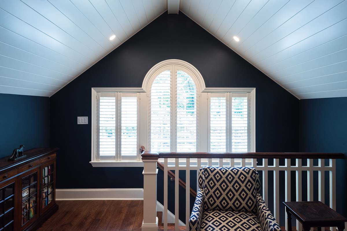 Cozy loft with a dark blue vaulted ceiling, white railing, and a patterned chair, featuring a large arched window and hardwood floors.